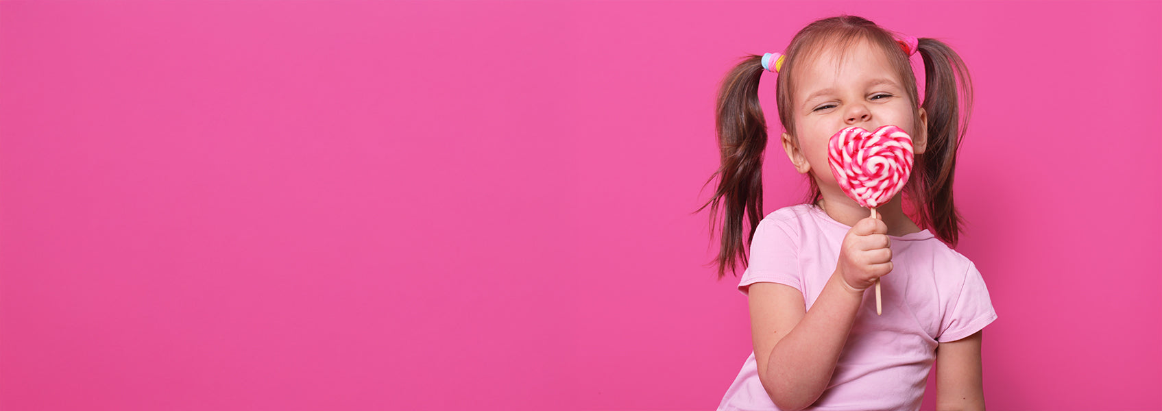 A young girl with pigtails wearing a light pink shirt smiles while holding a large pink and white heart-shaped lollipop in front of her mouth, against a bright pink background.