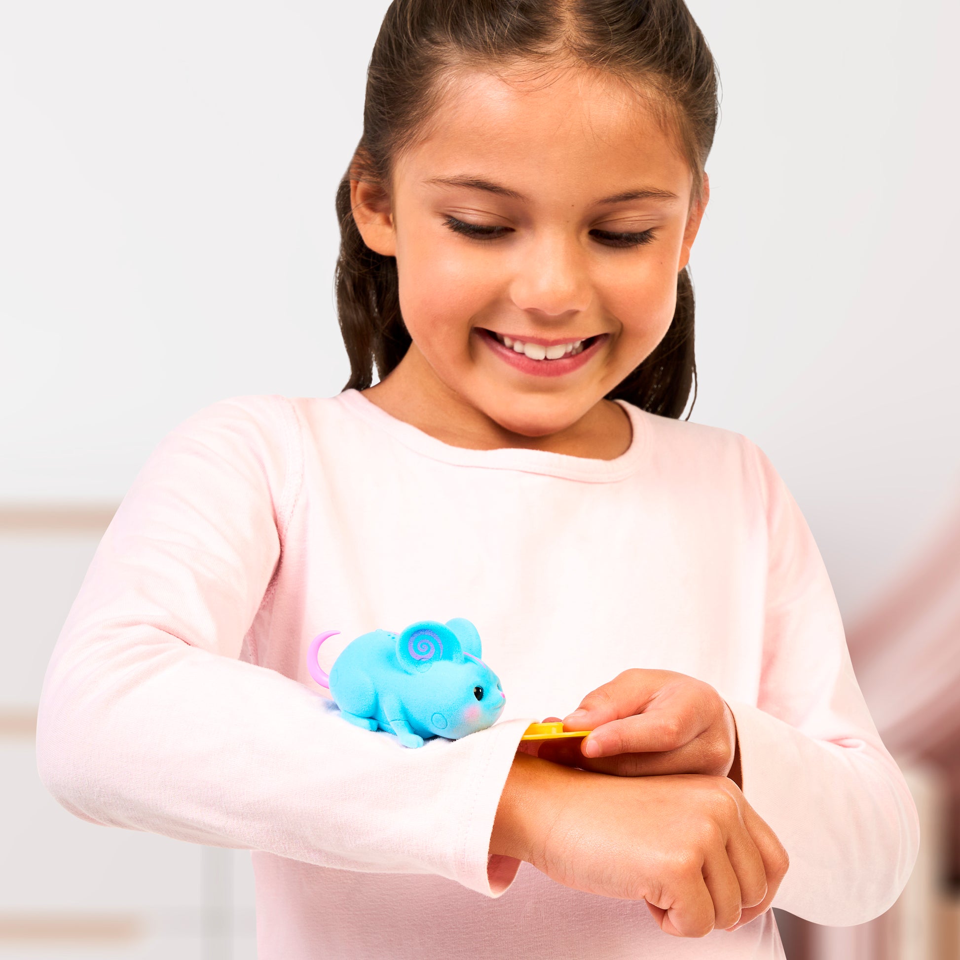 A young girl in a light pink long-sleeve shirt smiles as she controls a blue mouse from the Little Live Pets Needees Single Pack on her arm with a yellow button-like device.