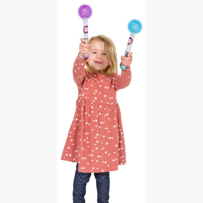A young child in a pink dress with white hearts smiles as she holds up two vibrant spiky sensory balls on sticks, resembling the Astroverse Cosmic Ray Light-Up Wand by Astroverse, against a white background.