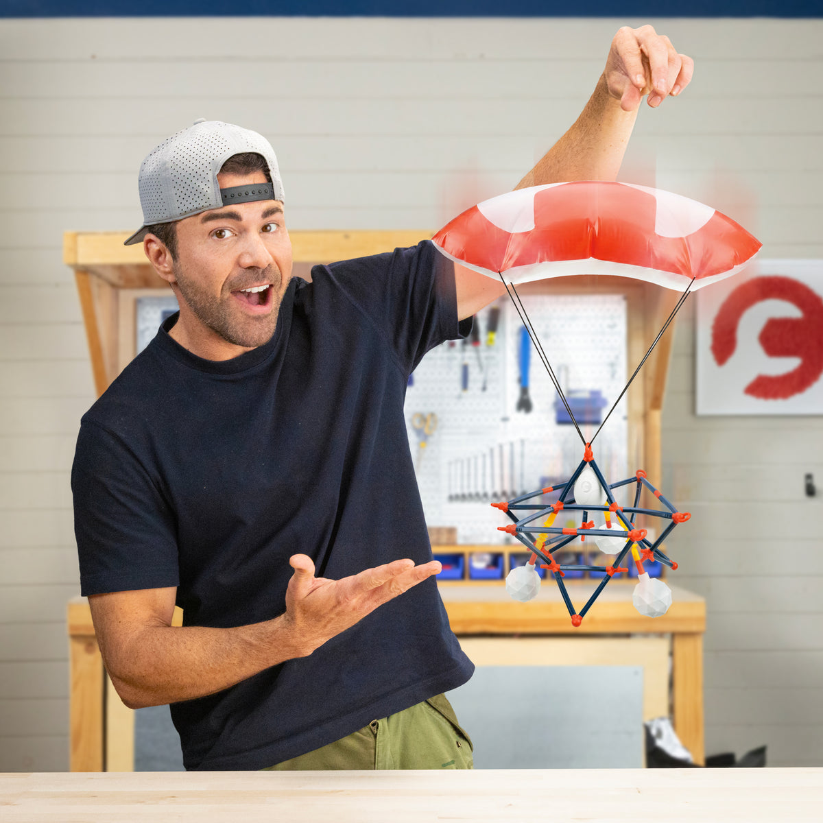 A man in a backwards cap smiles and gestures to a homemade parachute craft on strings, reminiscent of the Crunch Labs Crunch Kit Skyfall Egg Drop by Moose Toys, set in a workshop with tools on a pegboard in the background—the perfect creative space for experimenting with STEM kits from Crunch Labs.