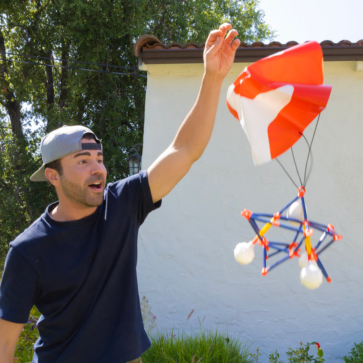 A man wearing a backward cap and dark shirt releases the Moose Toys Crunch Labs Crunch Kit Skyfall Egg Drop with its geometric parachute outdoors on a sunny day, smiling excitedly as he takes on the Egg Drop Challenge.