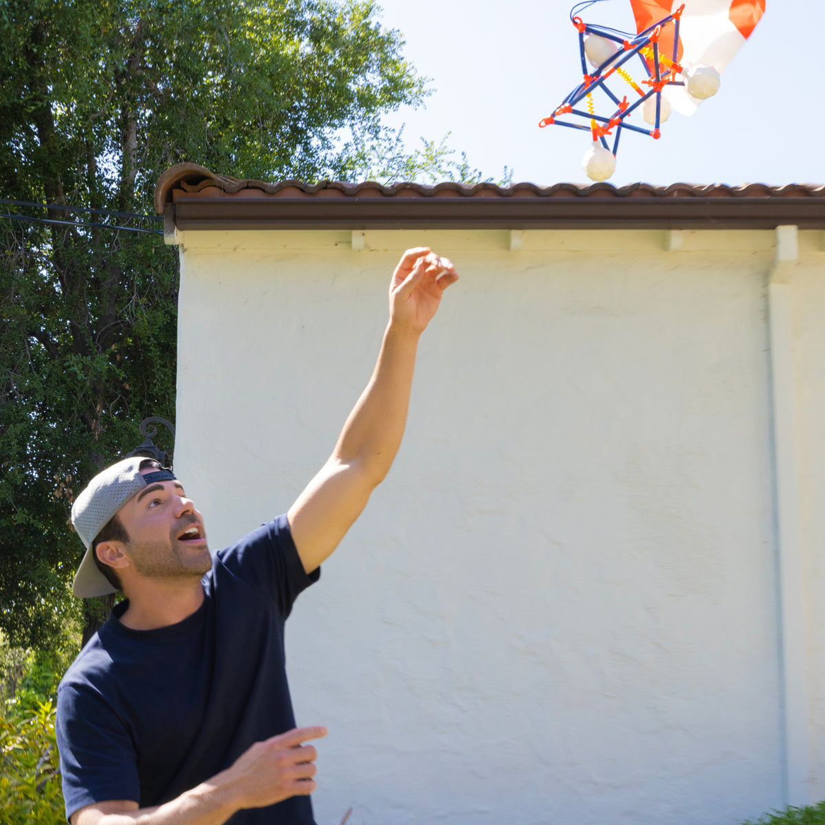 A person wearing a backwards cap reaches up excitedly toward a colorful, geometric object—like the Crunch Labs Crunch Kit Skyfall Egg Drop from Moose Toys—flying in the air near a house and trees.