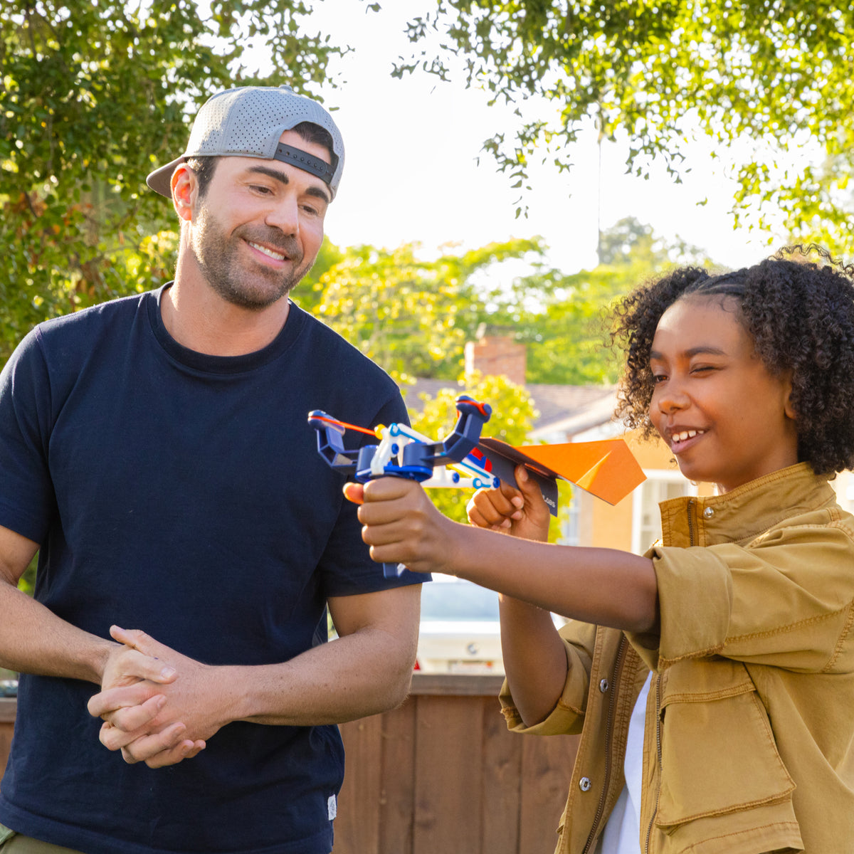 A smiling man watches as a child aims the Crunch Labs Crunch Kit Turbo Trick Flyer by Moose Toys outdoors, evoking inventive Mark Rober moments. Trees and a wooden fence are in the background.