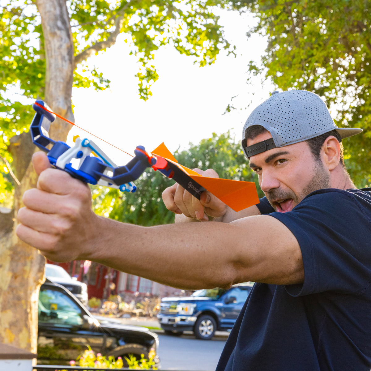 A man in a backward gray cap aims an orange arrow with a toy bow outdoors, standing on a street lined with trees and parked cars—capturing the inventive energy of Mark Rober and Moose Toys’ Crunch Lab Crunch Kits Single Pack Assortment.
