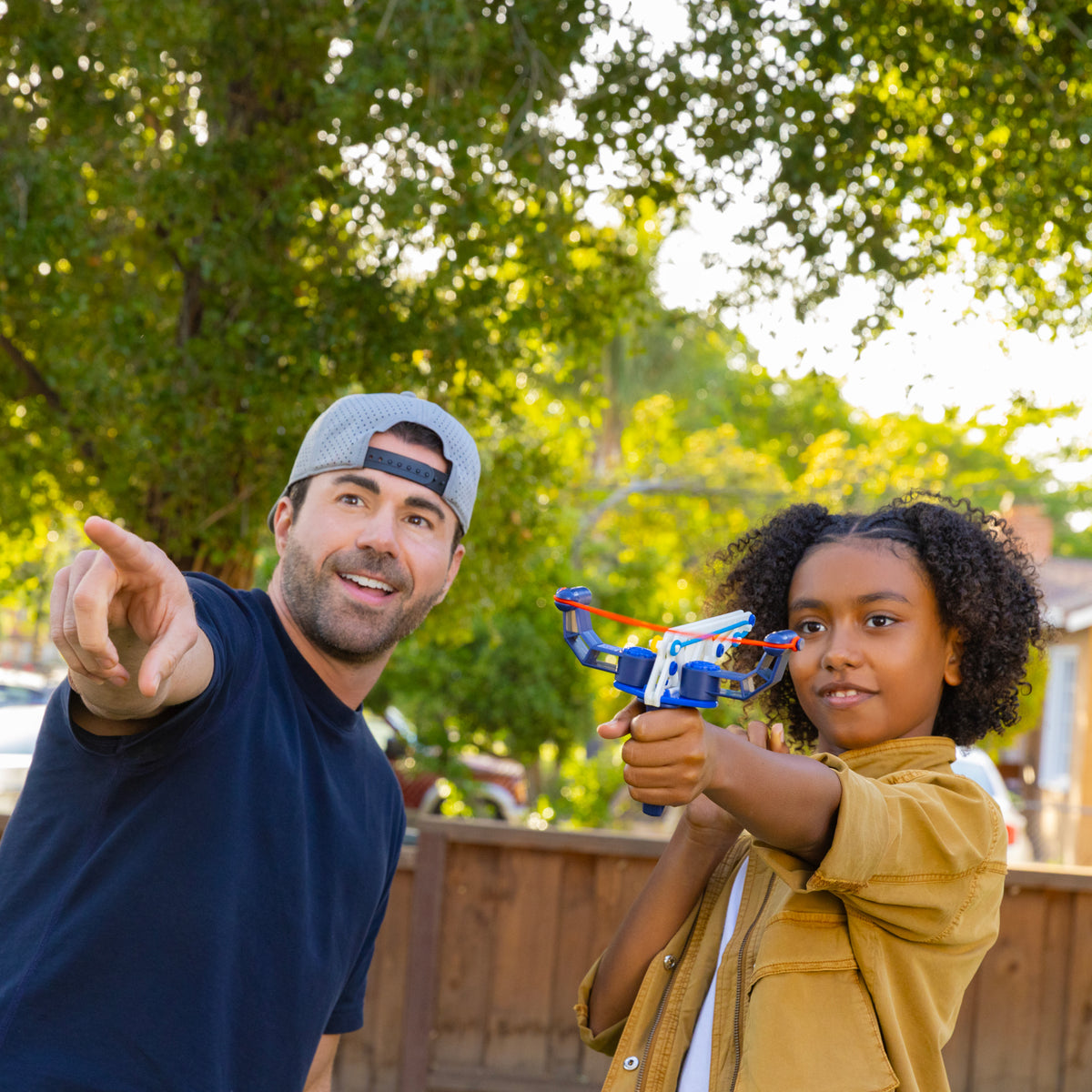 A smiling man in a backwards cap points while a child aims a Moose Toys Crunch Labs Crunch Kit Turbo Trick Flyer STEM kit. They are outside in a sunny yard with green trees in the background, channeling their inner Mark Rober as they explore hands-on science fun.