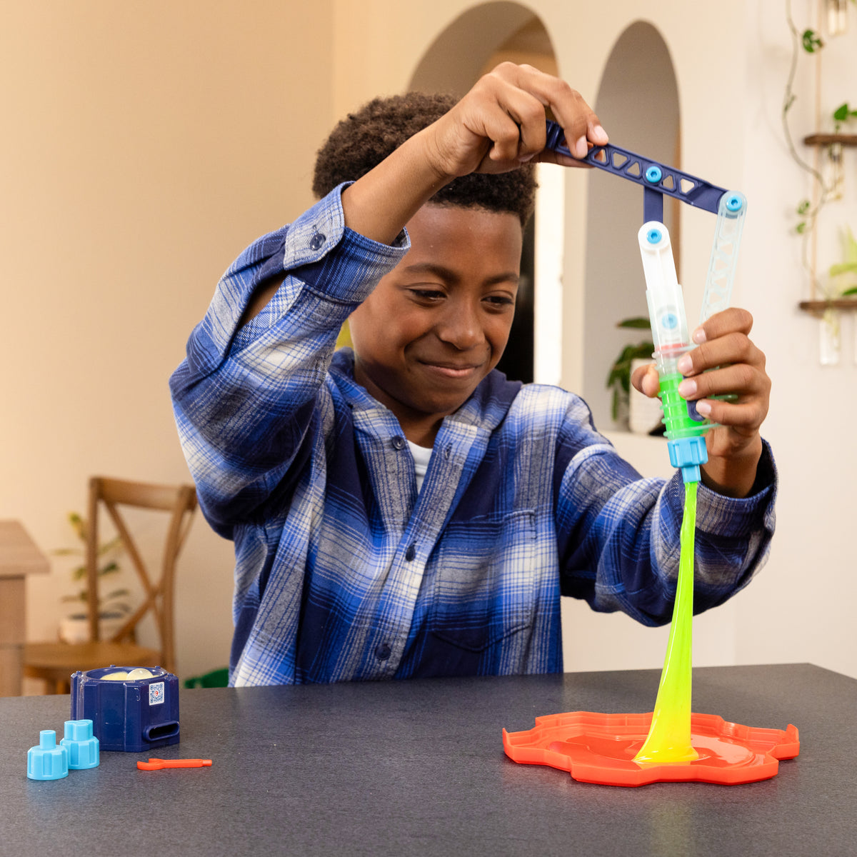 A boy in a blue plaid shirt smiles as he plays with the Moose Toys Crunch Lab Crunch Kit Slime Prank Lab, stretching bright yellow-green slime onto an orange tray at a table in a cozy, well-lit room.