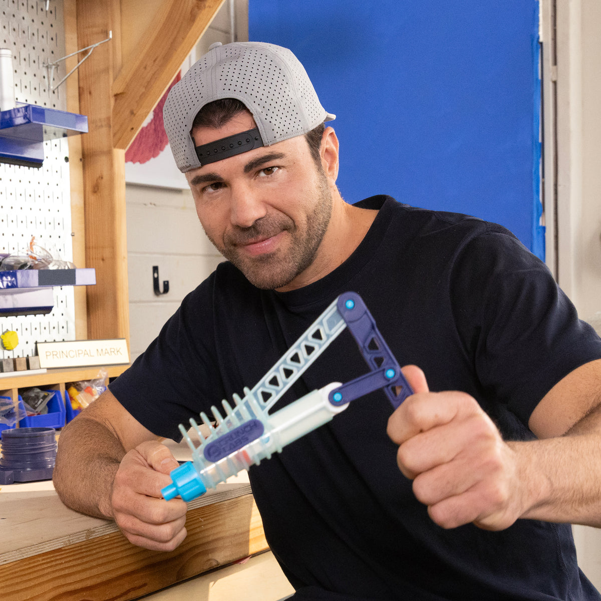 A man in a backward gray cap and black shirt smiles while holding a blue and white Moose Toys Crunch Lab Crunch Kit Slime Prank Lab in a workshop, with shelves and tools visible in the background.