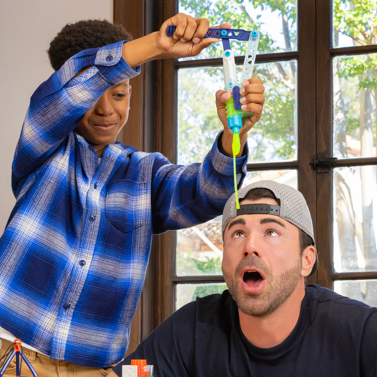 A smiling boy in a blue plaid shirt squirts liquid from a toy syringe above a surprised man in a backward gray cap inside a sunlit room, capturing the playful chaos of a Crunch Lab Crunch Kit Slime Prank Lab experiment by Moose Toys.