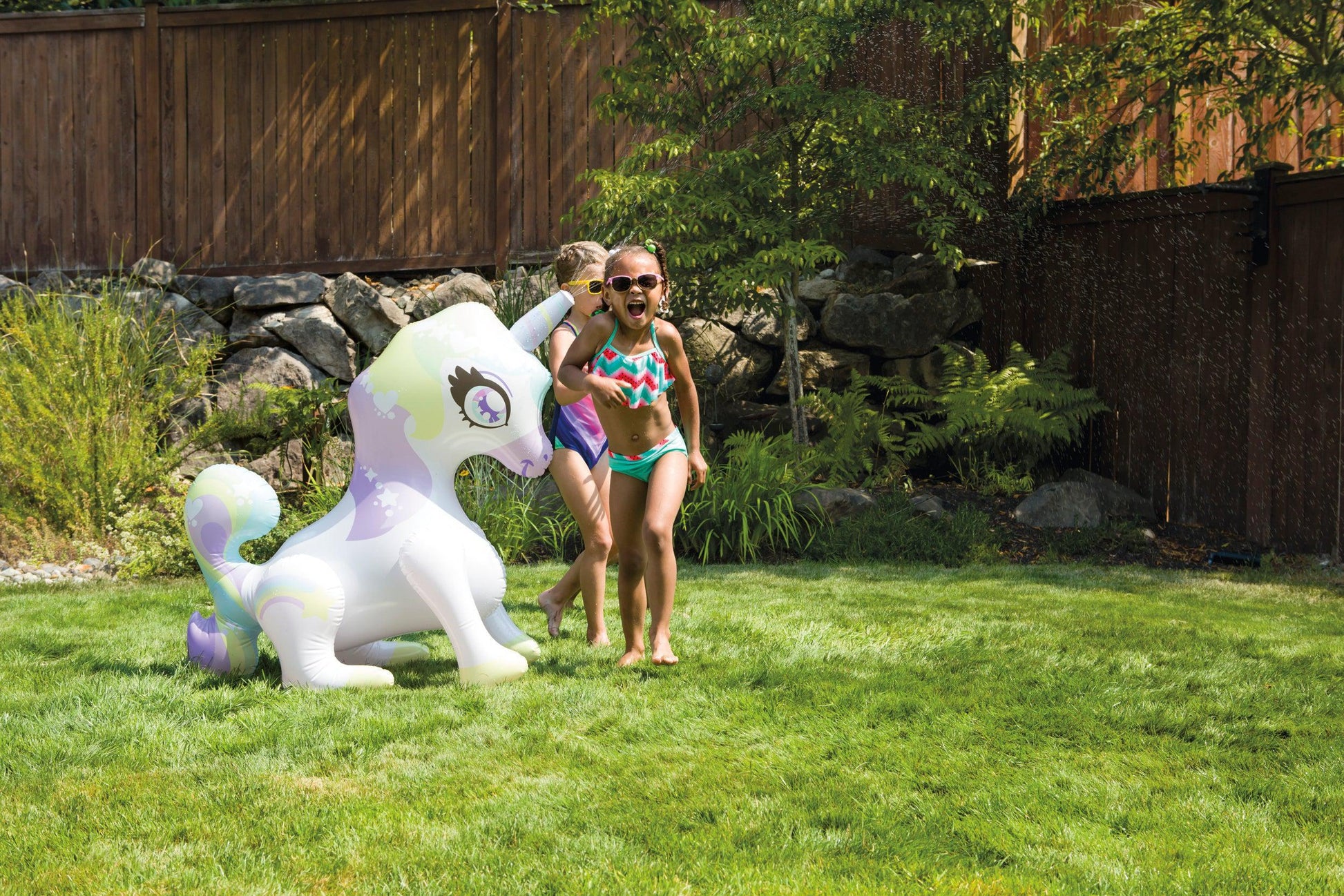 Two young girls in swimsuits laugh and play on the green grass, enjoying the refreshing spray of the Toysmith Sprinkler Buddies Mist-Ical Unicorn Sprinkler under the sunny sky, with water sparkling all around.