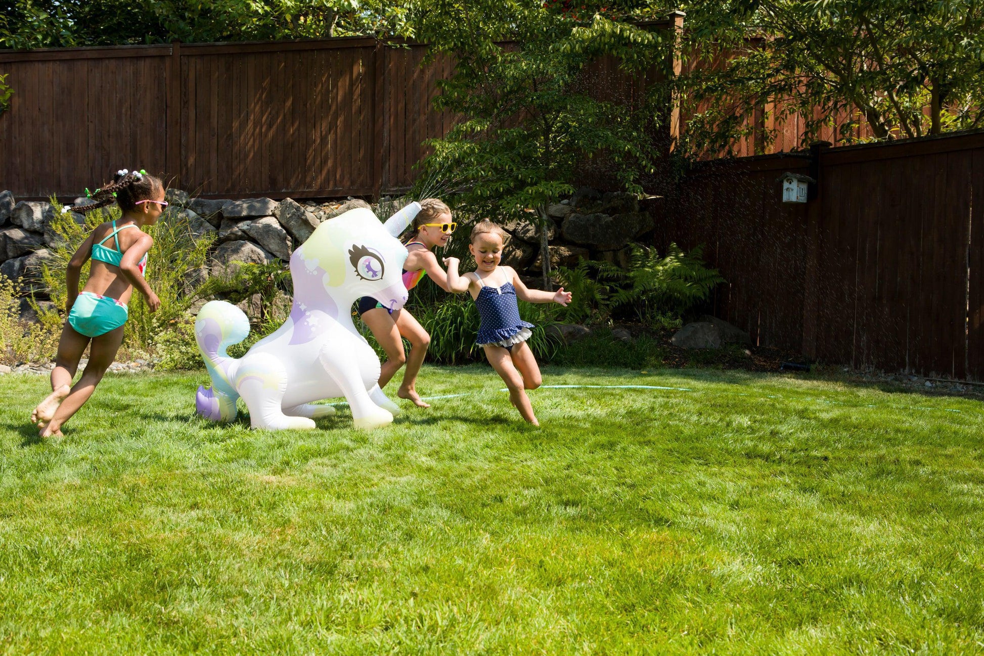 Three young children in swimsuits run and play on green grass as water sprays from the Toysmith Sprinkler Buddies Mist-Ical Unicorn Sprinkler in a sunny backyard, with a wooden fence and trees in the background.