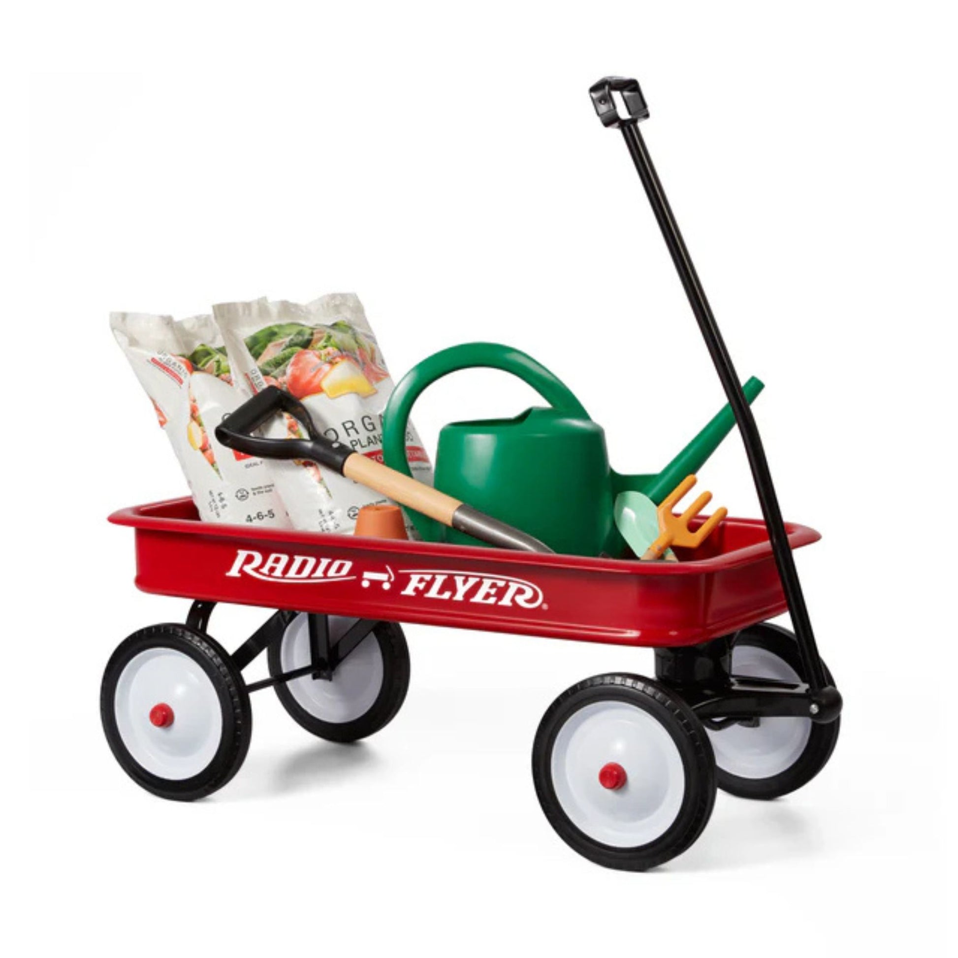 The Radio Flyer Classic Red Wagon by Radio Flyer holds gardening tools like seed bags, a green watering can, trowel, rake, and shovel. It features four wheels and a black pull handle for easy transport.
