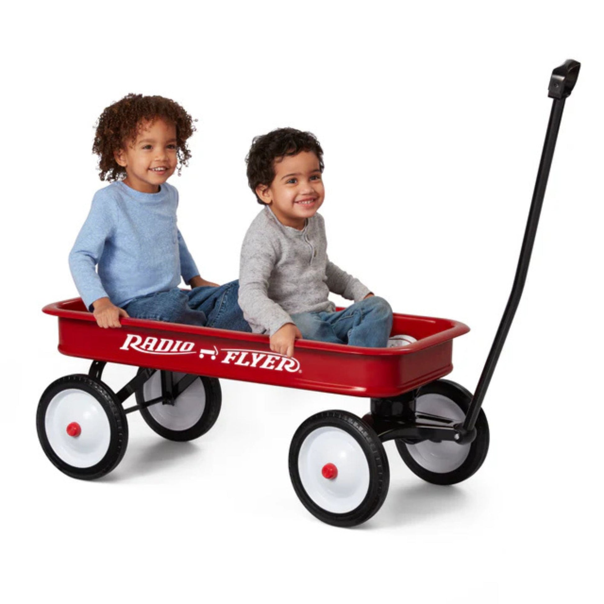 Two young children smile while sitting in a Radio Flyer Classic Red Wagon by Radio Flyer, featuring four large wheels and a black pull handle, set against a white background.