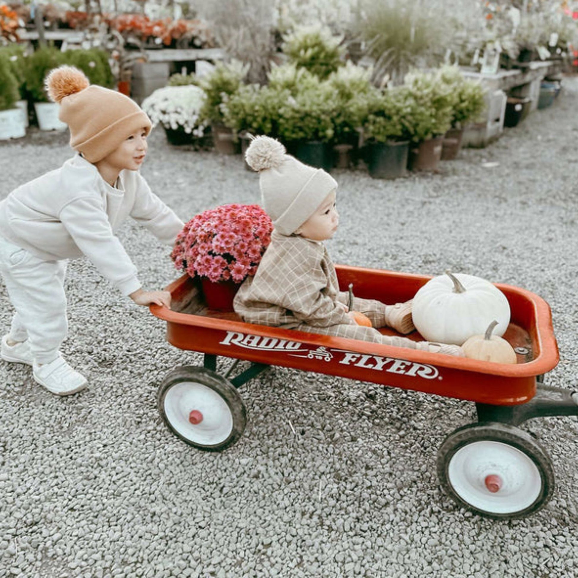 Two young children in knit hats enjoy the outdoors at a garden center as one pushes the other in a Radio Flyer Classic Red Wagon by Radio Flyer, which is filled with pink flowers and white pumpkins.