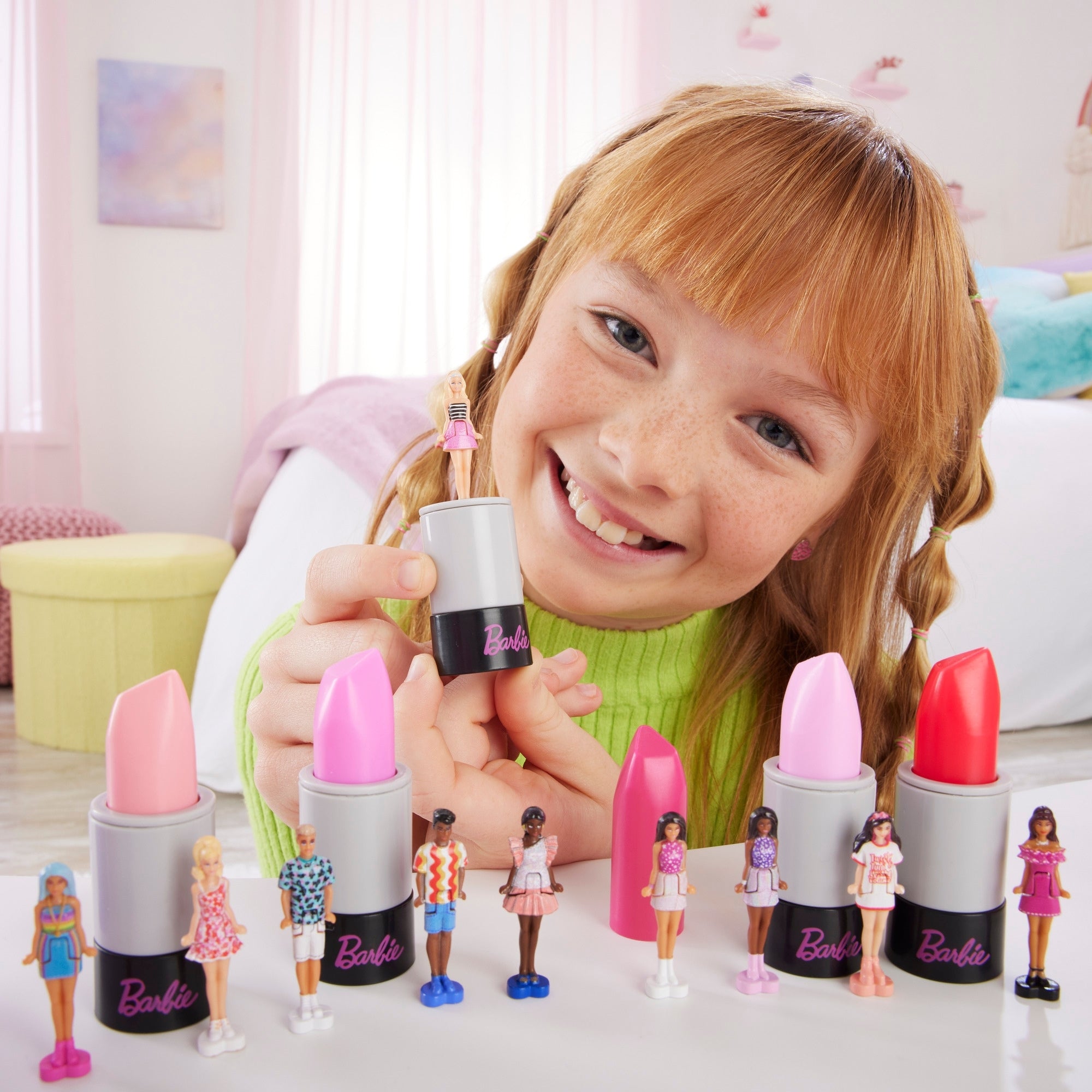 A smiling child holds a pink toy lipstick with a Mattel Barbie Mini Barbieland Doll on top. Several toy lipsticks and collectible dolls from the Mini Barbieland series, each sporting unique outfits, are arranged on the table before her.