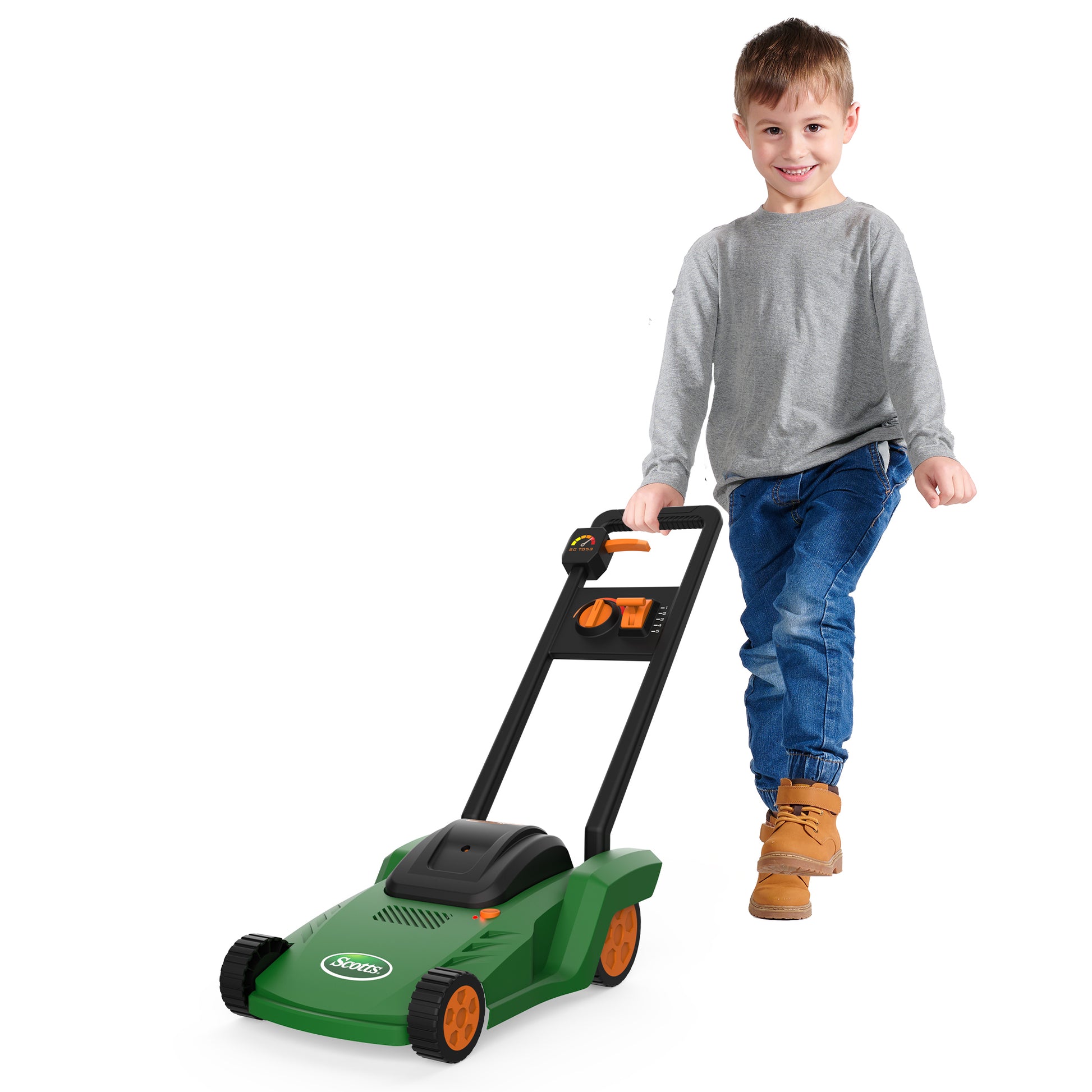 A young boy smiles as he pushes the Red Toolbox Scotts Water Squirting Lawnmower, featuring a green body and orange wheels, while wearing a gray long-sleeve shirt, blue jeans, and tan boots against a white background.