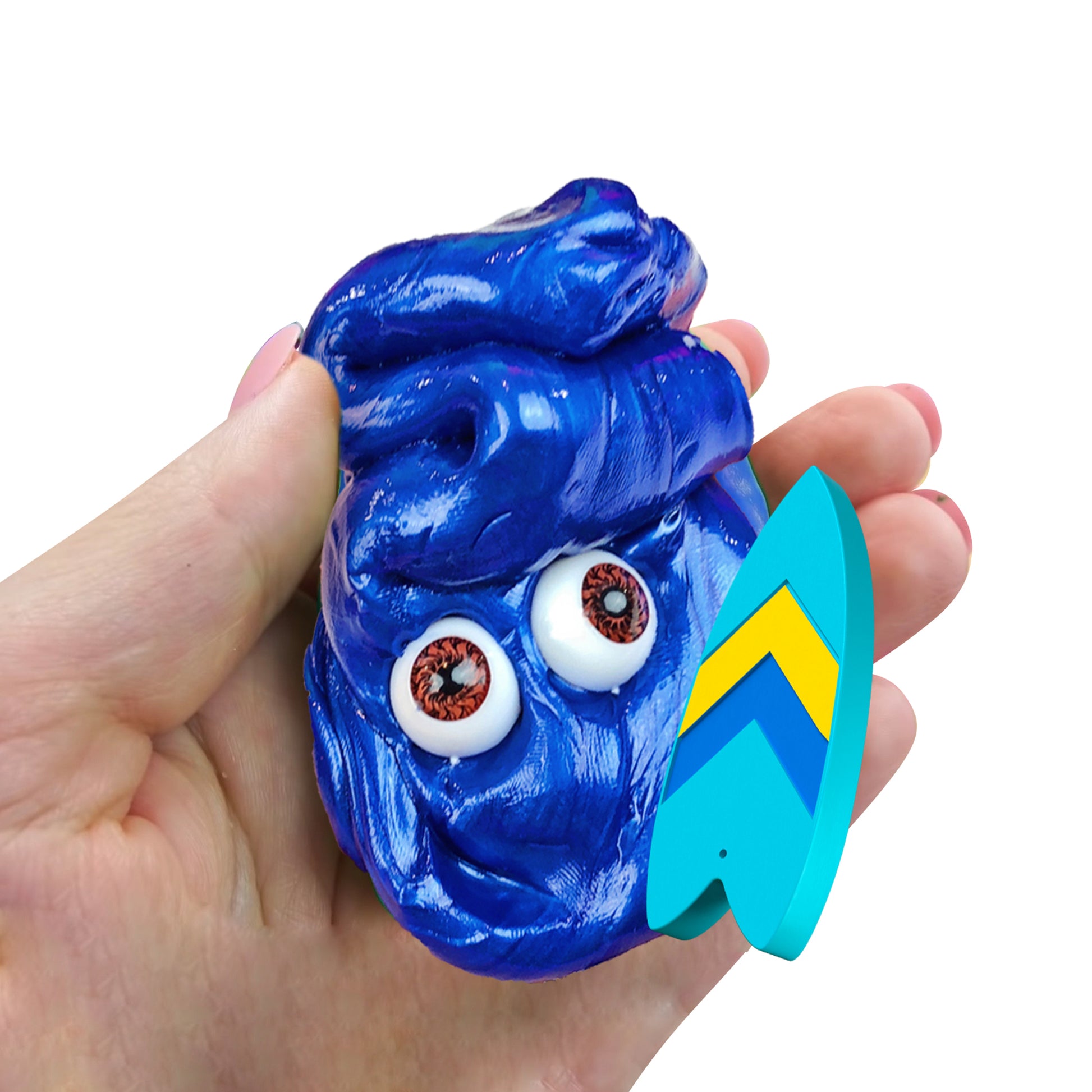 A hand holds a shiny blue Doctor Squish Putty Buddies Asst. 21ct CDU with two fake eyes and a tiny blue-and-yellow surfboard, shaping the putty into a fun, quirky face.