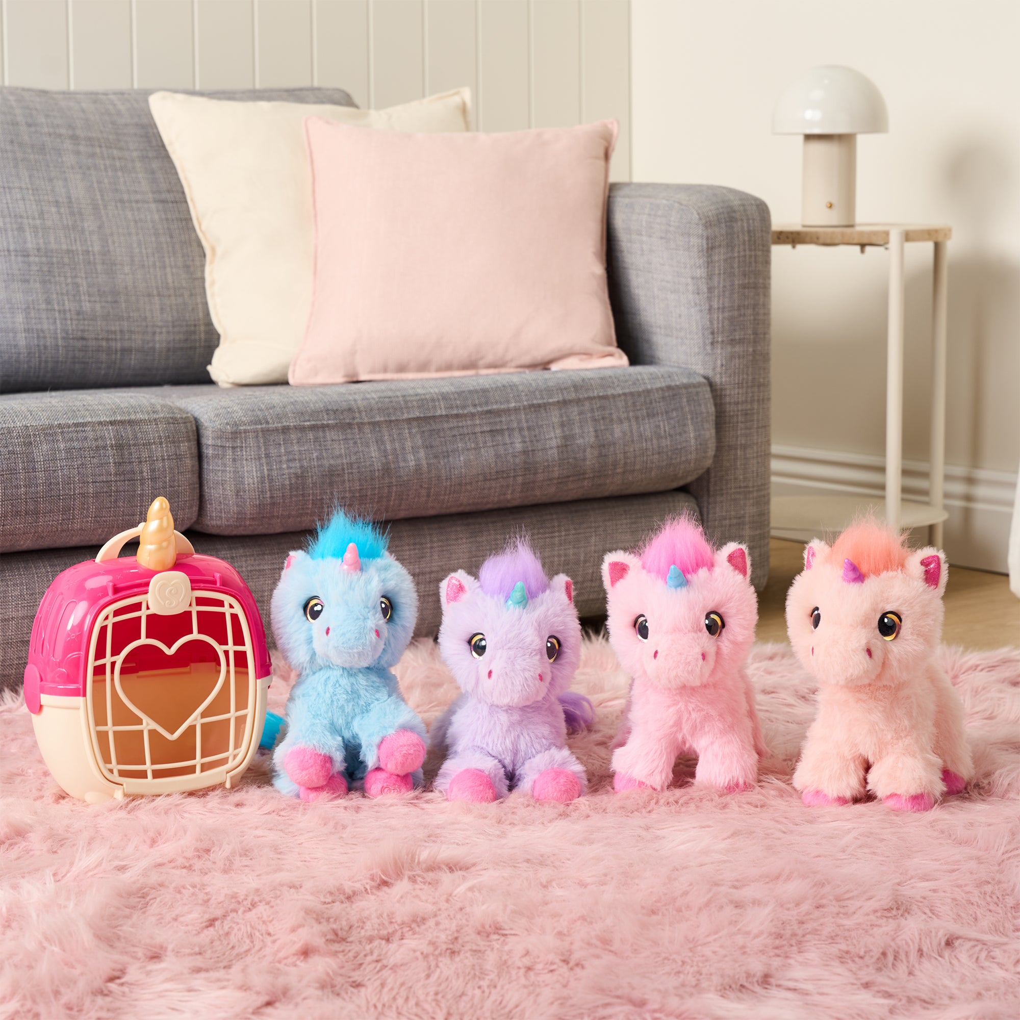 Four colorful unicorn plush toys, including the ScruffaLuvs S3 Unicorn from Moose Toys, are lined up on a pink fuzzy rug by a gray sofa. Nearby, a unicorn carrier rests with a cushion and a white lamp visible in the background.