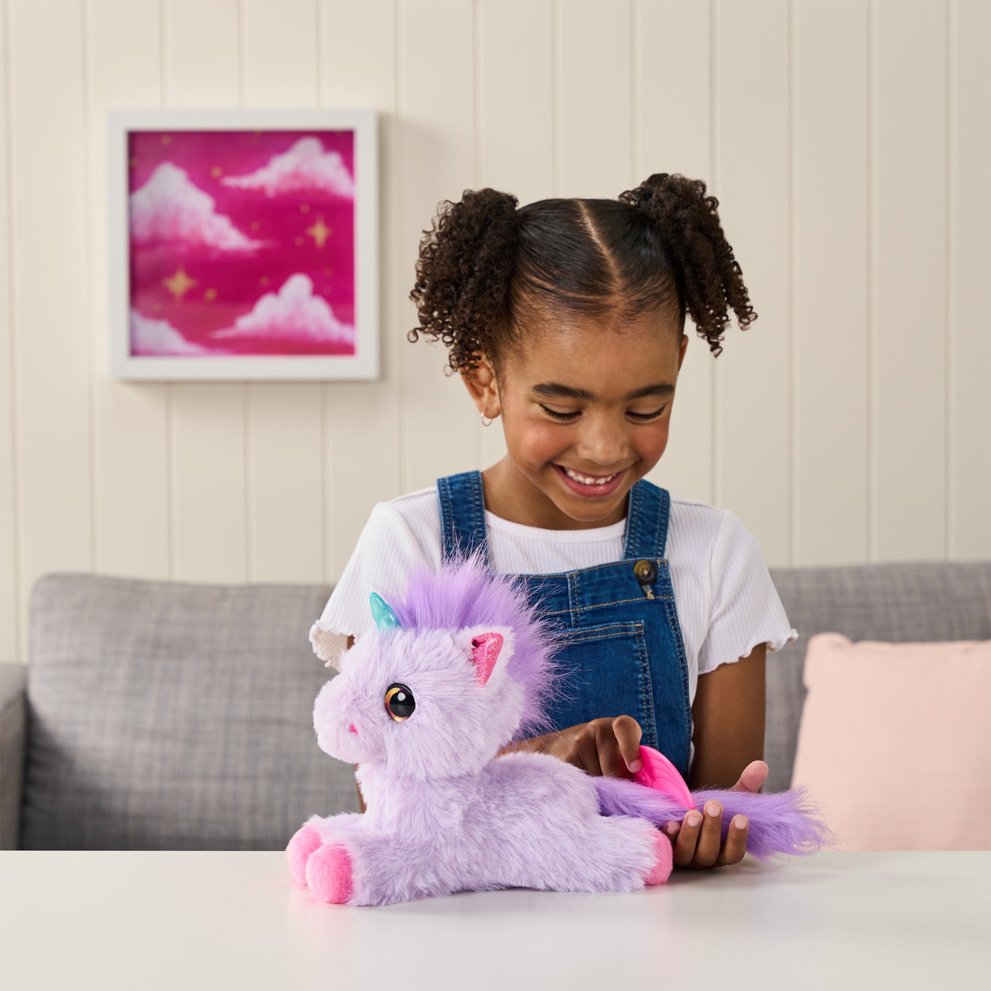A young girl smiles while playing with the Moose Toys ScruffaLuvs S3 Unicorn plush at a white table, sitting on a gray couch with a framed pink unicorn-themed picture on the wall behind her.