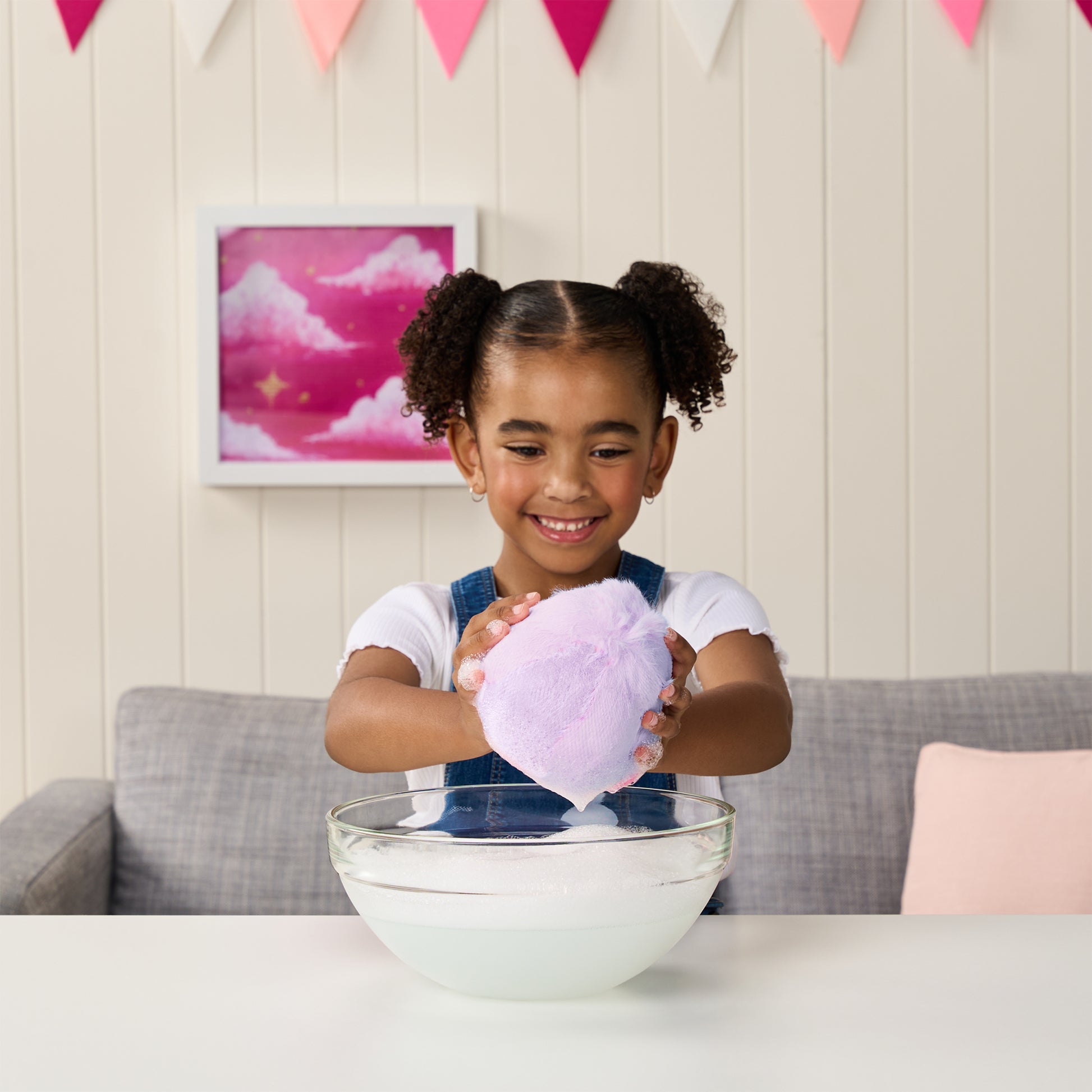 A smiling young girl squeezes a large purple bath bomb over a bowl at a white table, with her Moose Toys ScruffaLuvs S3 Unicorn plush by her side and pink bunting plus framed art in the background.