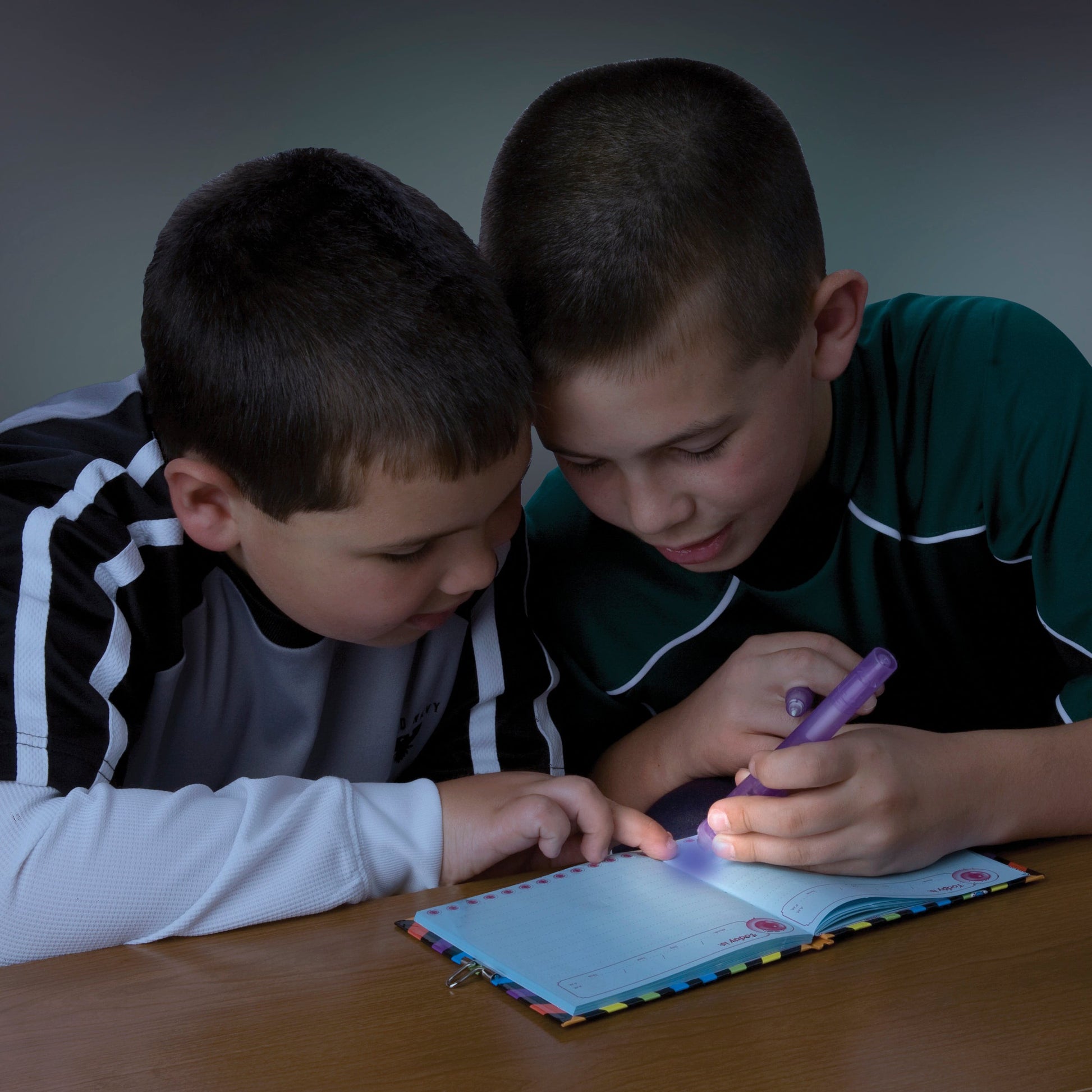 Two boys sit at a table, closely examining a notebook as one uses the IQ 2 in 1 Invisible Writer to create secret messages, which are revealed with the pen’s built-in UV light in the dimly lit room.