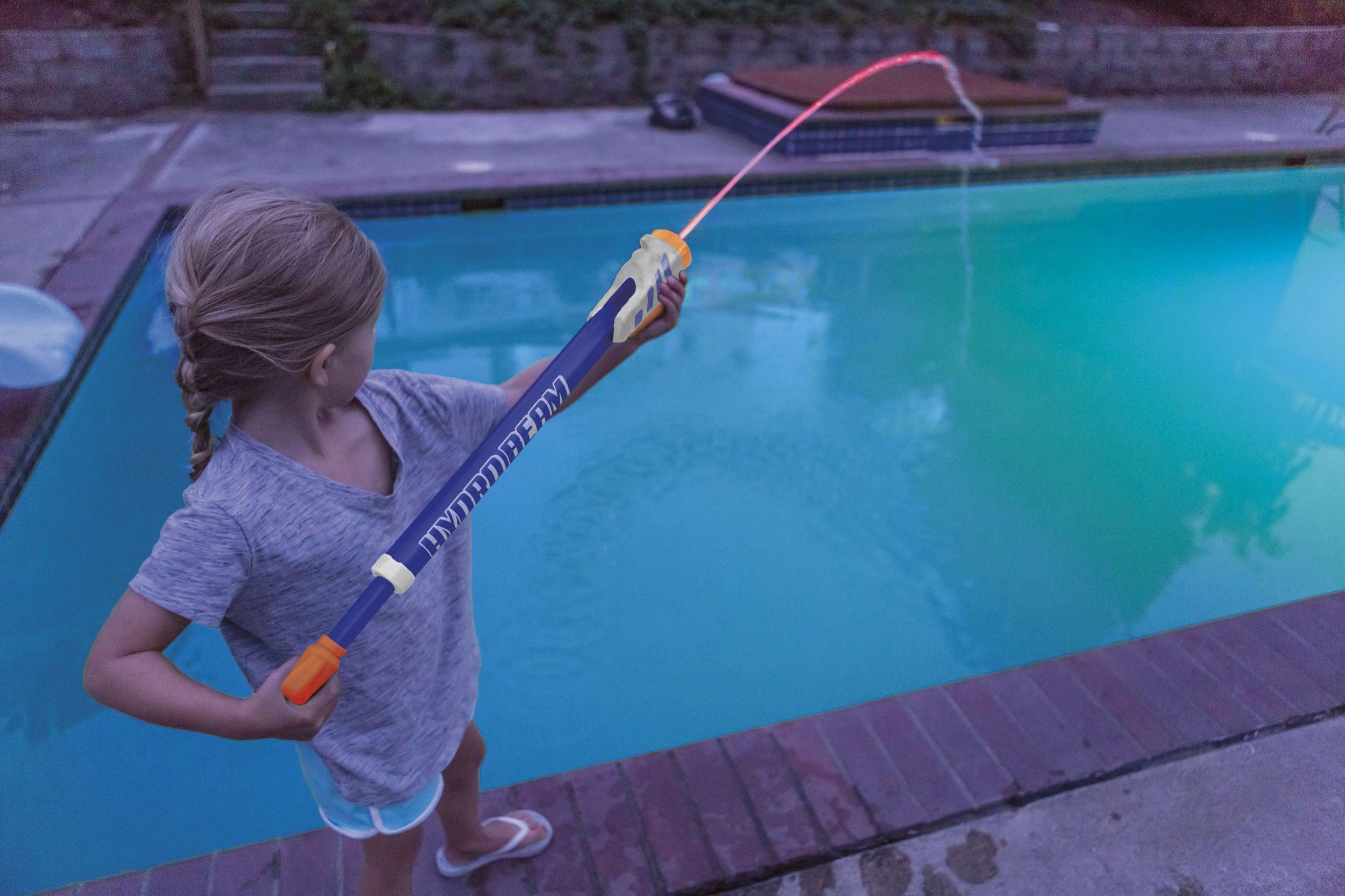 A young girl with braided hair stands by a swimming pool in the evening, spraying water into the pool using the GO! Play Hydro Beam Water Launcher from GO! Play.