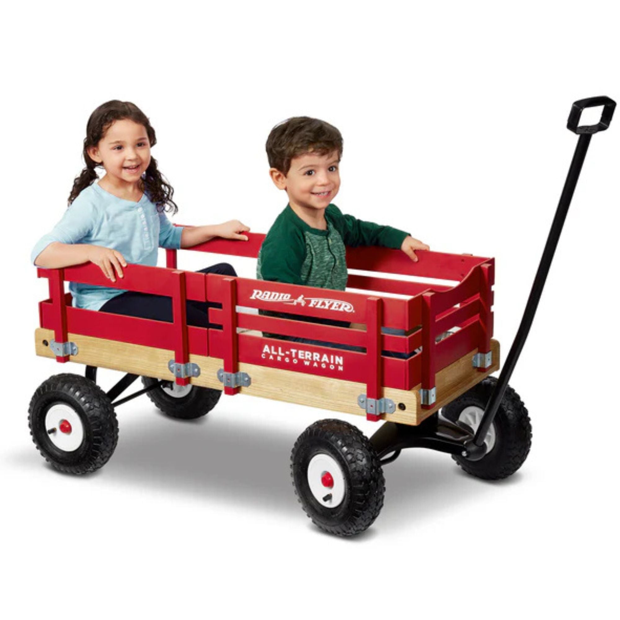 Two young children smile as they sit in a Radio Flyer All Terrain Cargo Wagon, featuring black all-terrain wheels and a long black handle, by the iconic Radio Flyer brand, set against a plain white background.