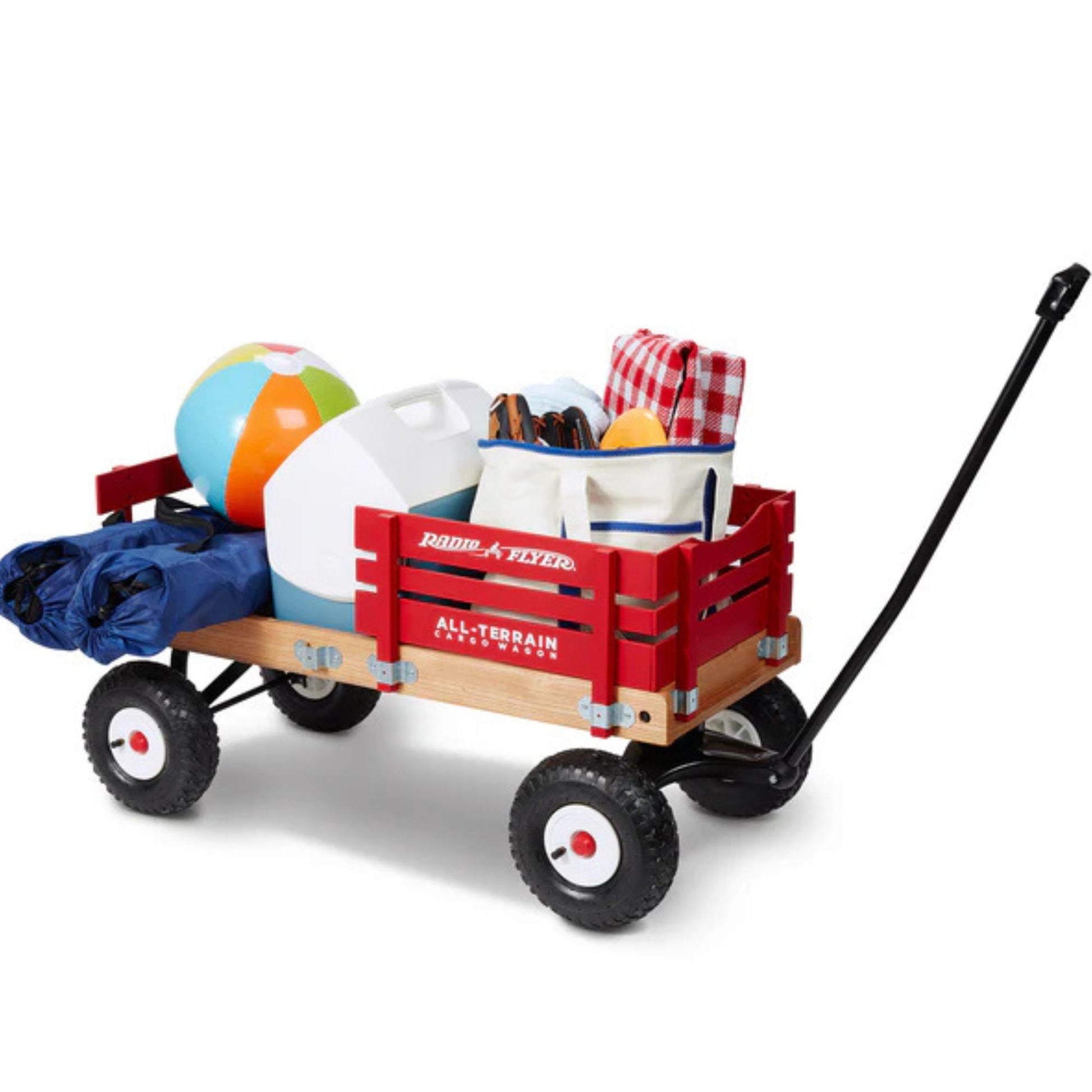 A Radio Flyer All Terrain Cargo Wagon, by Radio Flyer, packed with a beach ball, folded picnic blanket, cooler, jug, and outdoor essentials; features a black handle and large wheels for all terrains.