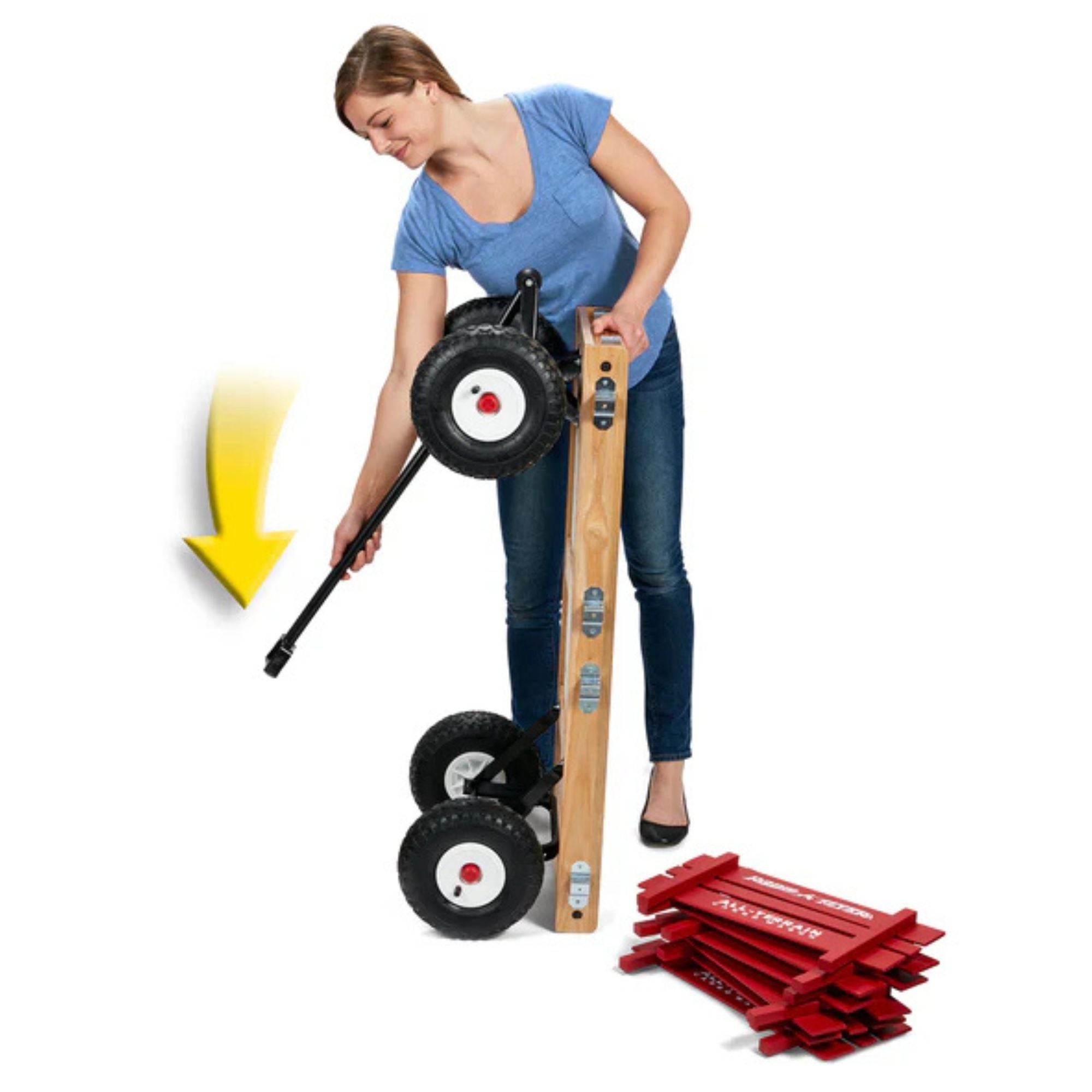 A woman attaches a wheel to a Radio Flyer All Terrain Cargo Wagon with two wheels already on. An arrow highlights the wheel, and several red plastic leveling boards are stacked nearby on the floor.