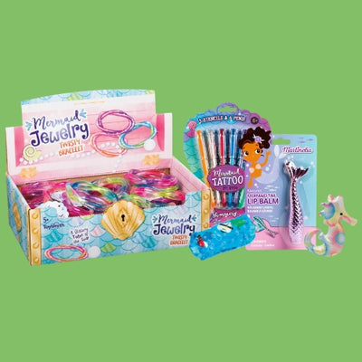 Colorful mermaid-themed toys and accessories including twist bracelets, temporary tattoos, lip balm shaped like a mermaid tail, and a small unicorn-mermaid figurine, displayed on a green background.