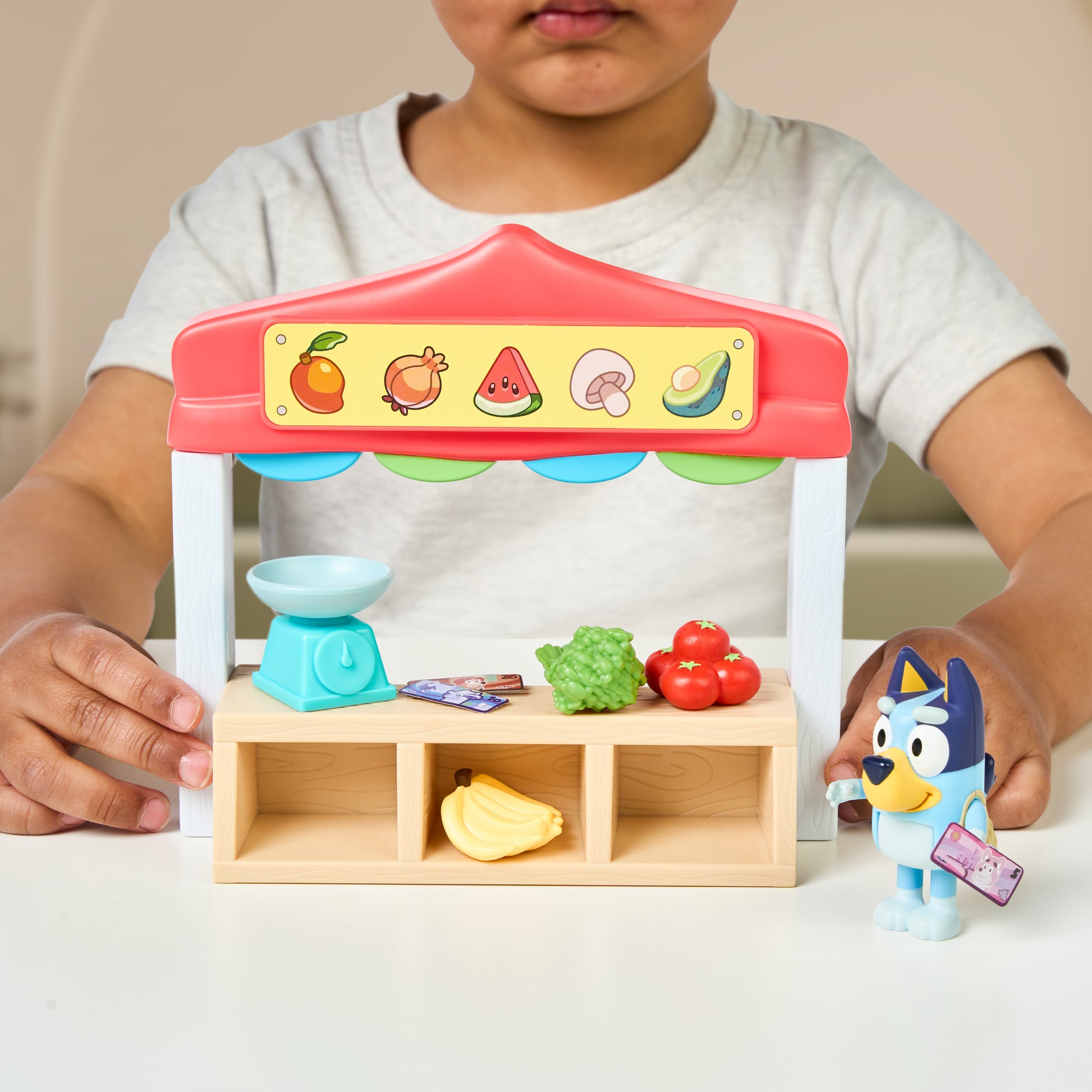 Child playing with a Bluey toy fruit stand and vegetables on a table.