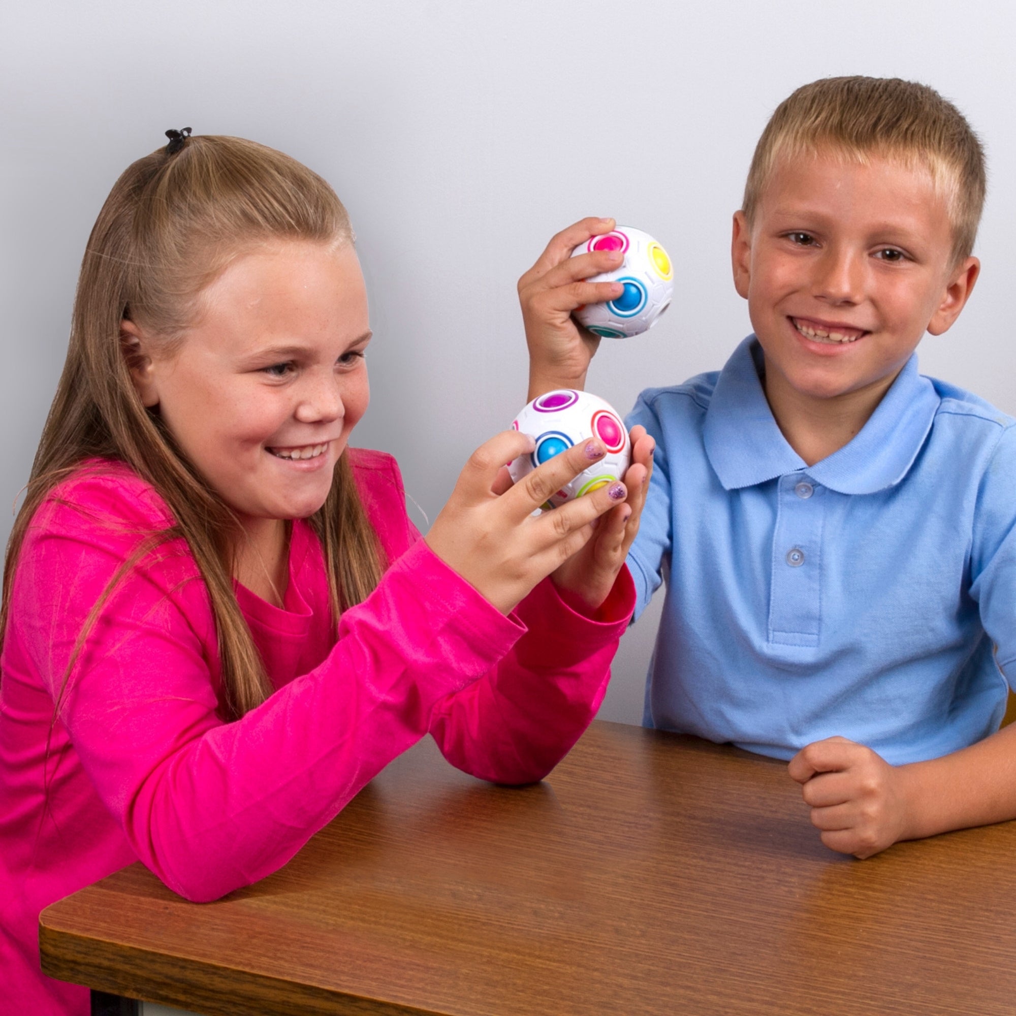 A girl in a pink shirt and a boy in a blue shirt smile at a table while playing with Duncan Color Shift Puzzle Balls, pressing the colorful buttons.