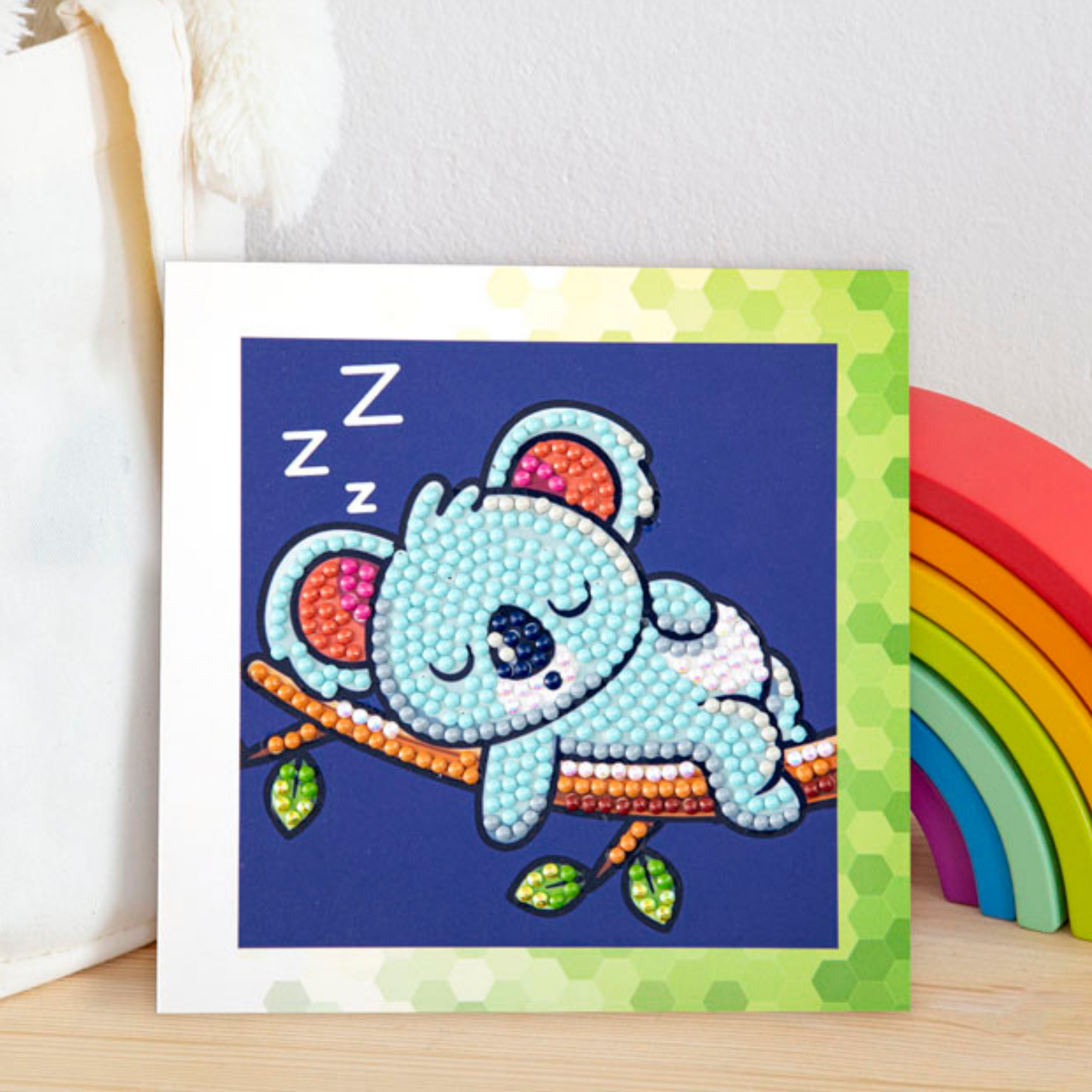 The Diamond Dotz Koala card showcases a cute blue koala sleeping on a branch with dot art accents and green geometric borders, accompanied by a vibrant rainbow.