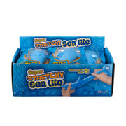 A blue Toysmith display box labeled "Super Stretchy Sand Filled Sea Life" holds stretchy blue sea life sensory toys in plastic packs. The box shows a hand stretching a toy, says Collect All 4!, and is for ages 3+.