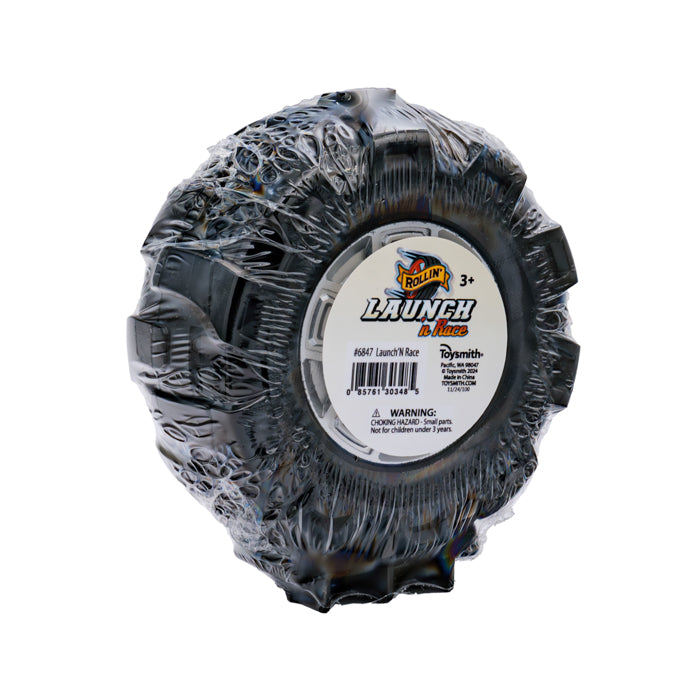 A black toy tire wrapped in plastic with a label reading "Rollin' Launch 'N Race" by Rollin'. Ideal for racing play, the label shows age 3+, Rollin' branding, and a choking hazard warning.
