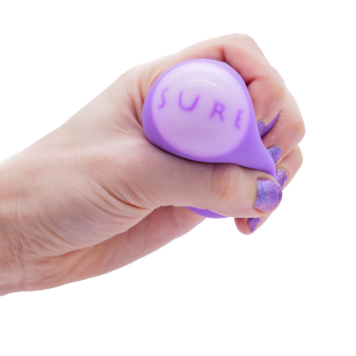 A hand with glittery purple nail polish squeezes a Toysmith Squeezy Fortune Ball—a purple squishy toy by Toysmith that reveals the word "SURE" when pressed. The background is white.