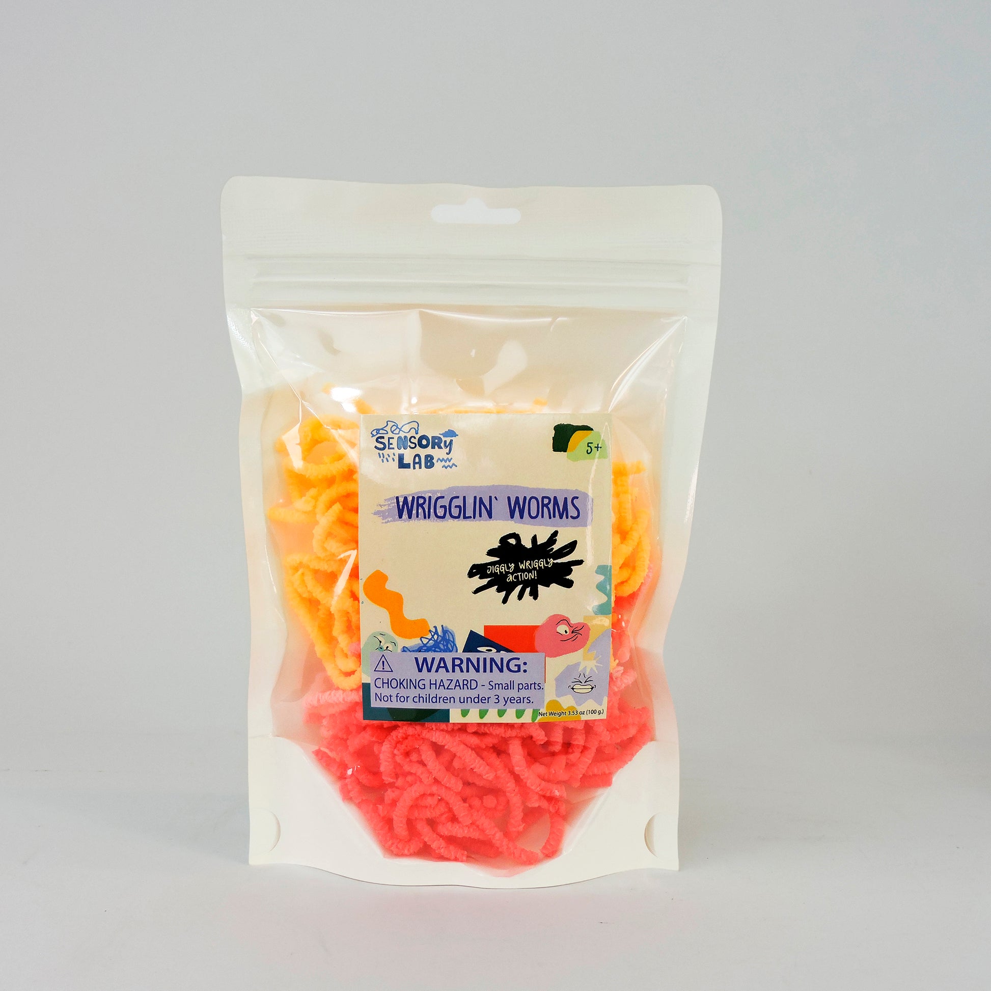 A clear resealable bag labeled Sensory Lab Wrigglin Worms contains orange and yellow worm-shaped sensory toys. The packaging warns of choking hazards and notes the product is not for children under 3 years.