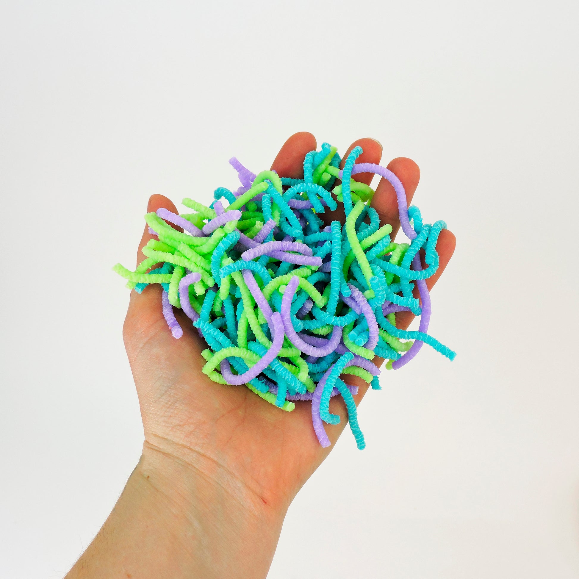 A hand holds a pile of Sensory Lab Wrigglin Worms—colorful fuzzy craft pipe cleaners in green, blue, and purple—against a plain white background.