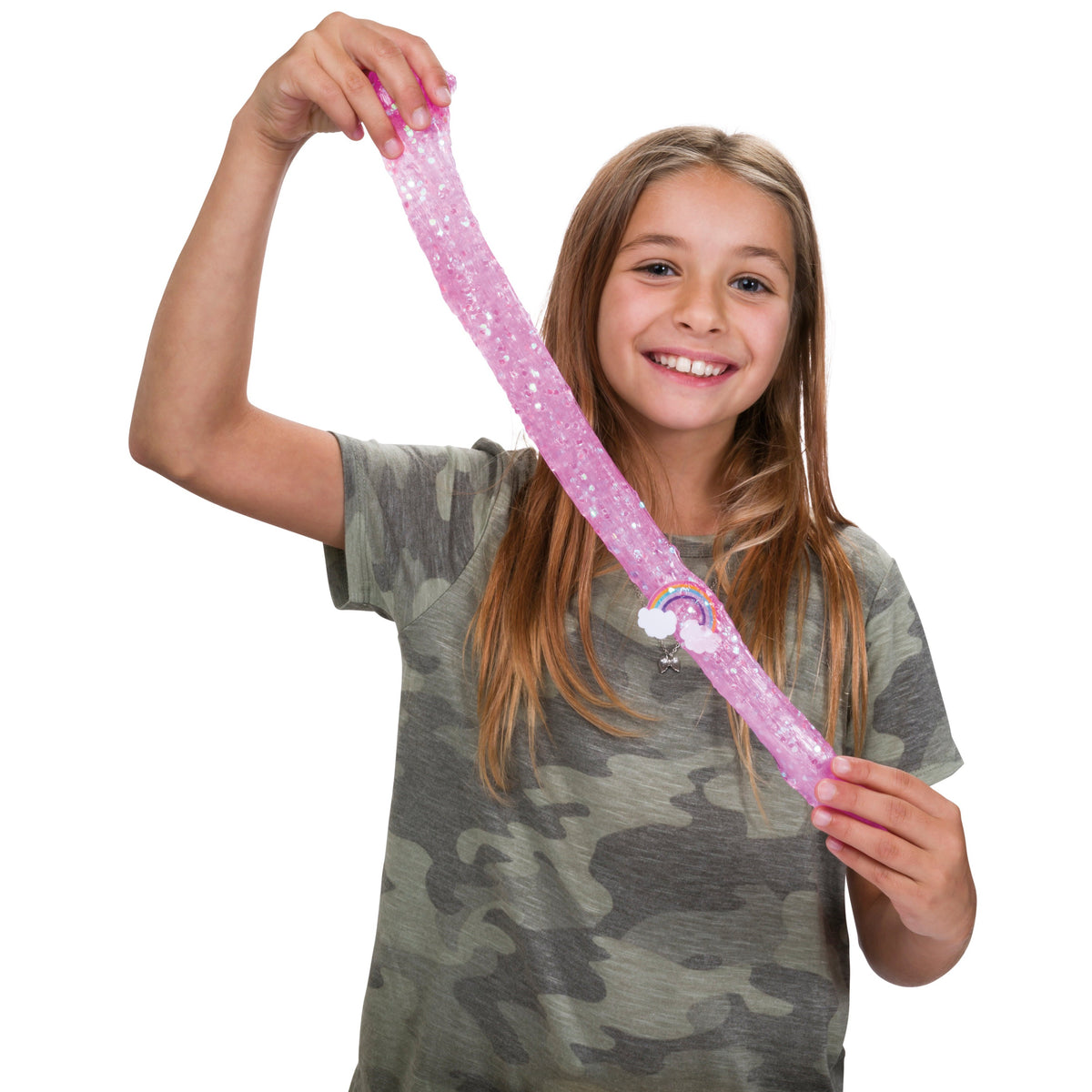 A smiling girl with long blonde hair wears a camouflage t-shirt as she stretches Toysmith Mix-Ins Slime Asst by Toysmith, a sparkly pink slime toy, between her hands against a white background.