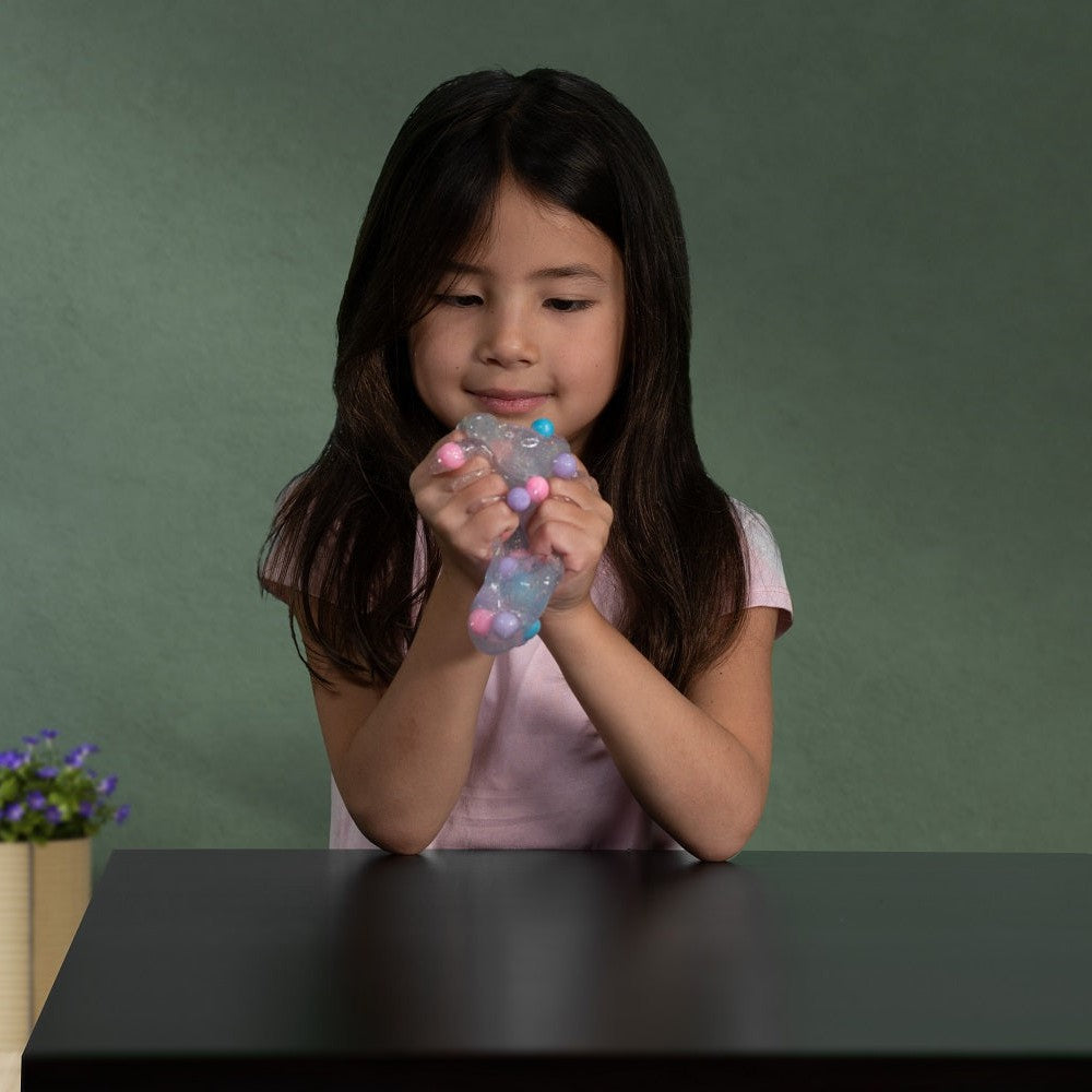 A young girl with long dark hair, in a pink shirt, smiles while squeezing Toysmith Polka Dot Slime at a dark table. The colorful sensory toy glimmers in her hands, with a small potted plant visible in the background.