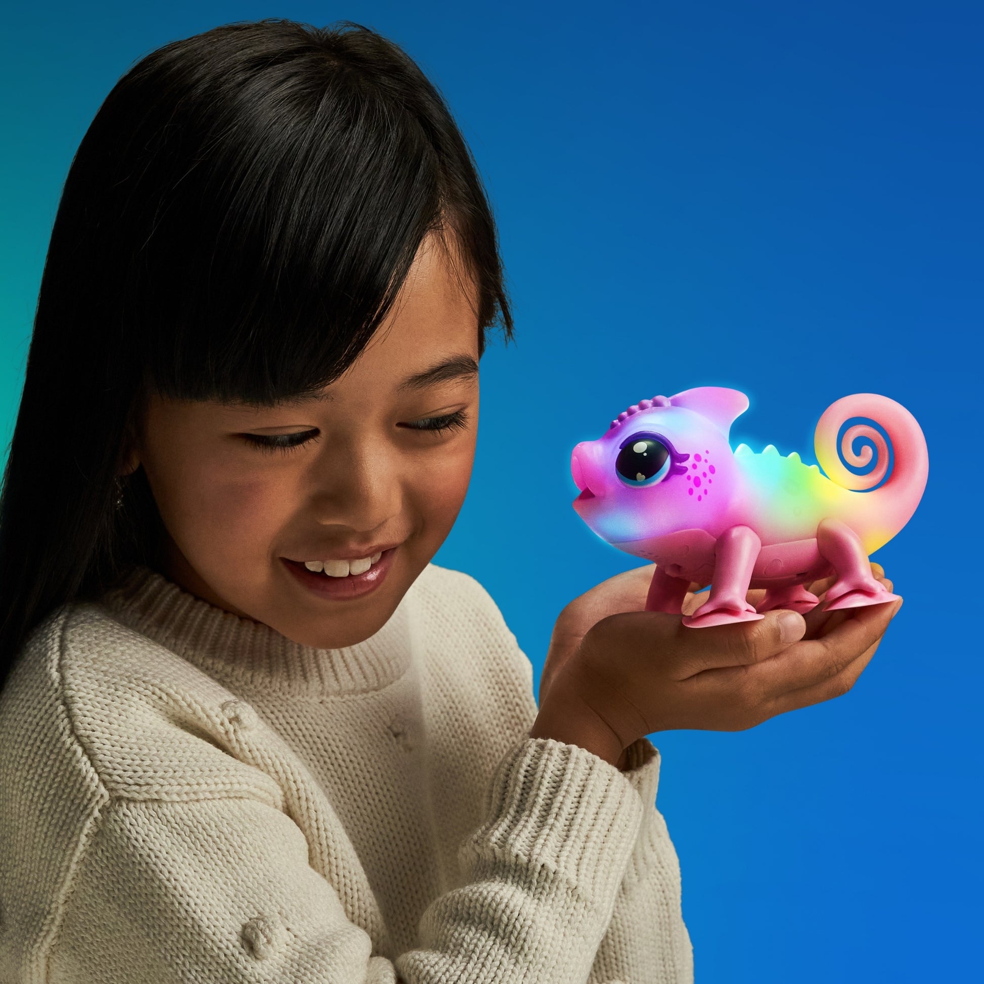 A smiling young girl in a white sweater holds the Moose Toys Little Live Pets S2 Lil' Chameleon, an interactive pet that glows with vibrant colors against a blue background.