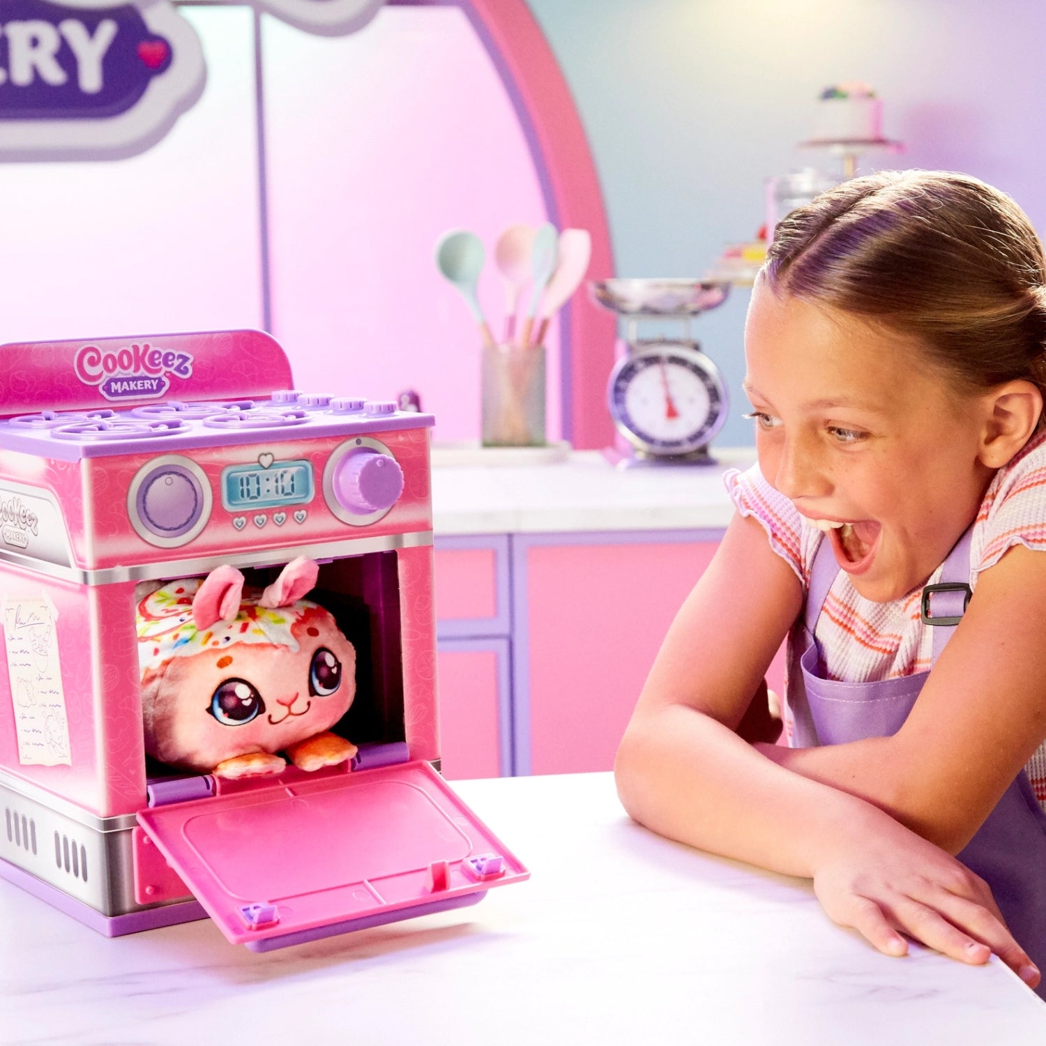 Child playing with a pink toy kitchen set, looking excitedly at a Cookeez toy.