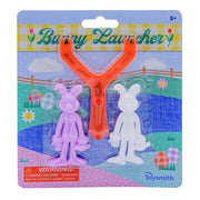 The Toysmith Bunny Launcher Easter basket toy includes an orange slingshot and two bunny figures, one purple and one white, displayed on a festive spring-themed background with a warning label.