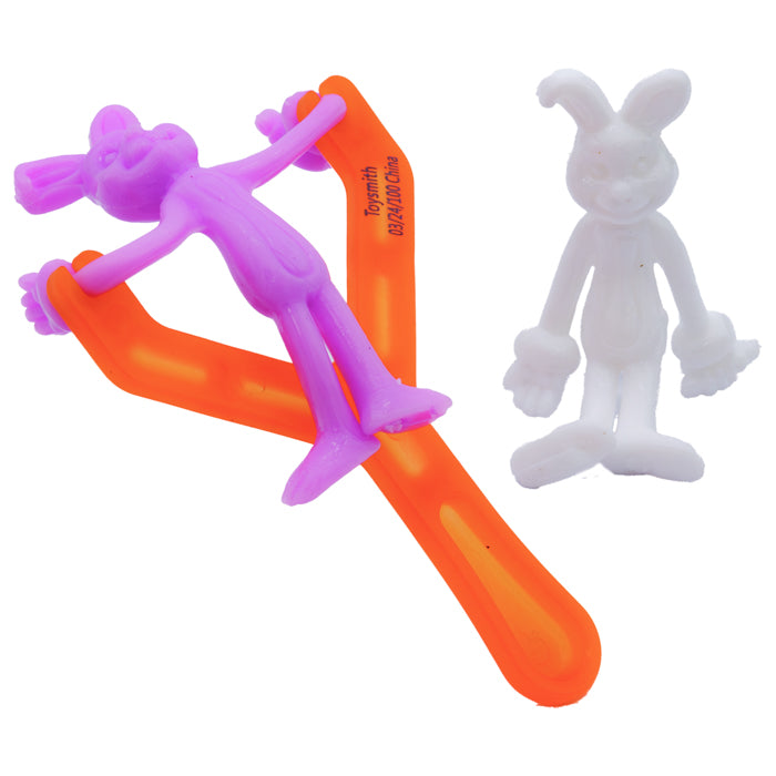 Two stretchy cartoon bunny toys—one purple, one white—are shown. The purple bunny is stretched across the Toysmith Bunny Launcher for springtime play, while the white bunny stands nearby. Perfect as an Easter basket toy!.