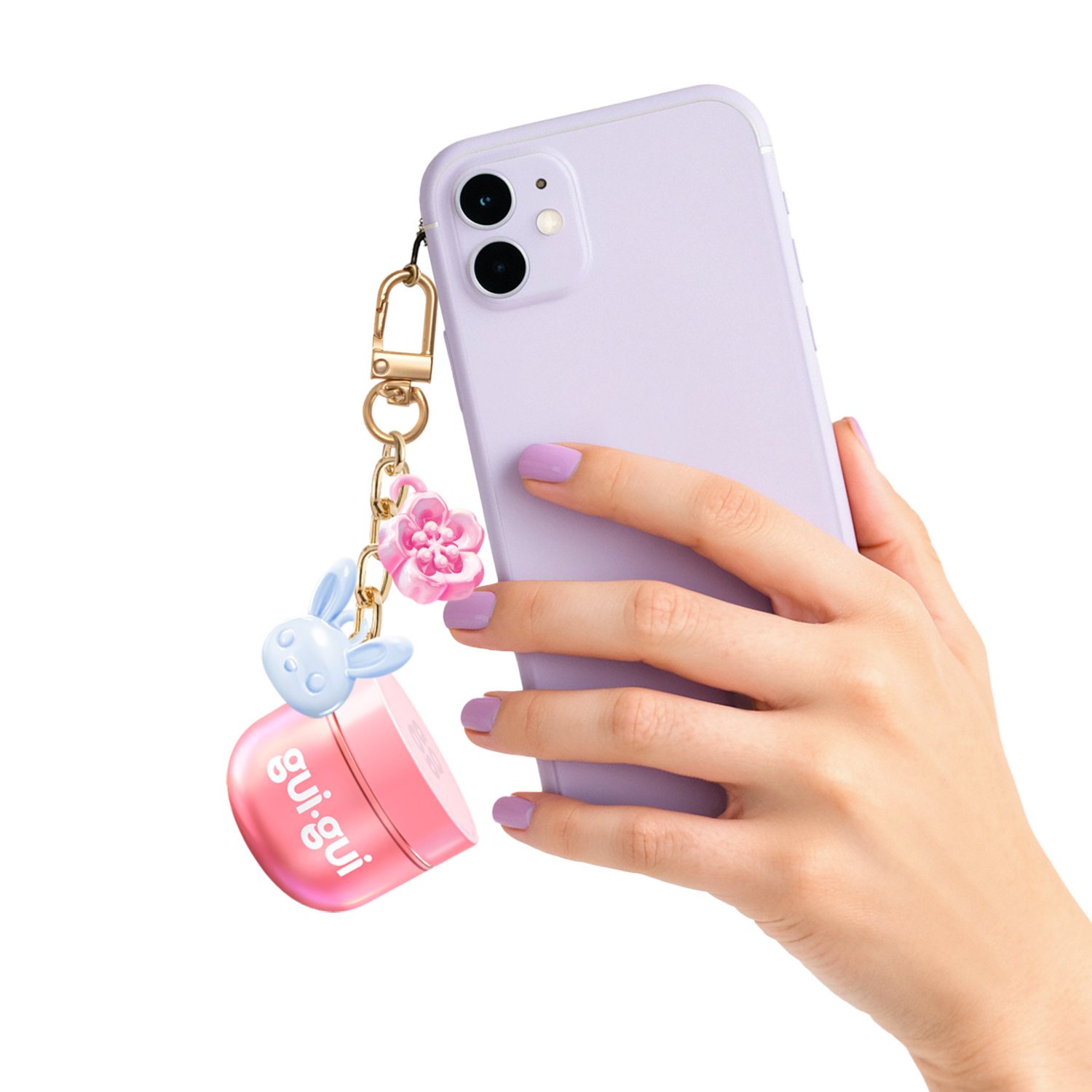A hand with lavender-painted nails holds a lavender smartphone with a keychain featuring a pink flower, a blue bunny charm, and the Moose Toys Gui Gui Mini Backpack Charm Slime.