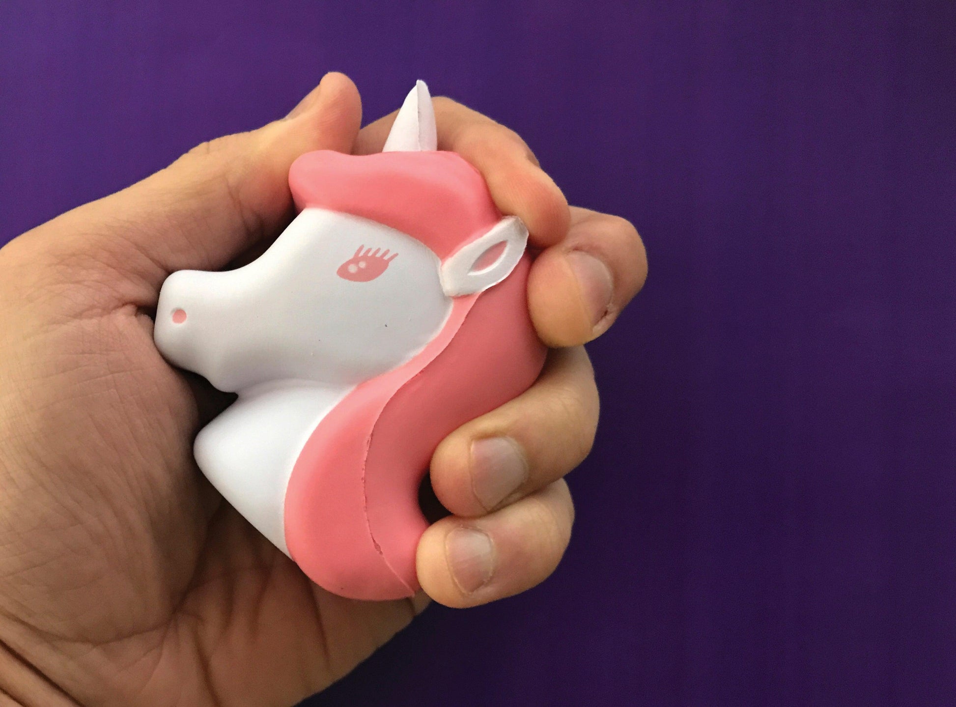 A hand squeezes The Source Unicorn Stress Ball, featuring a pink mane and closed eye, made of squishy foam, against a solid purple background.
