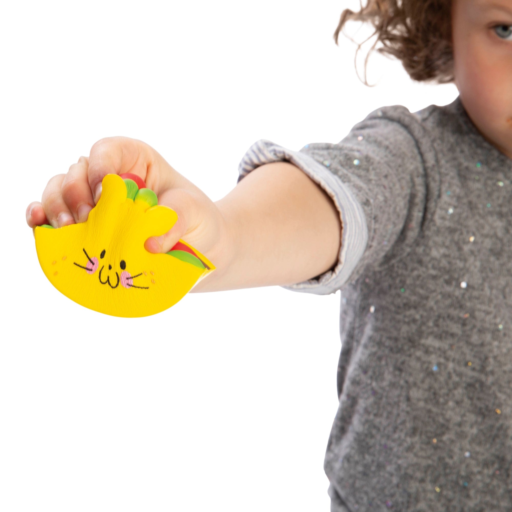 A child in a gray shirt holds out the Toysmith Taco Cat Squishy, a yellow cat-shaped toy with cute facial features and pink cheeks by Toysmith, against a white background.