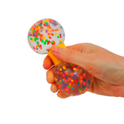 A hand squeezes the Sensory Lab Squeez-E Swirls, a clear stress ball filled with colorful confetti-like dots, making the top bulge against a plain white background.