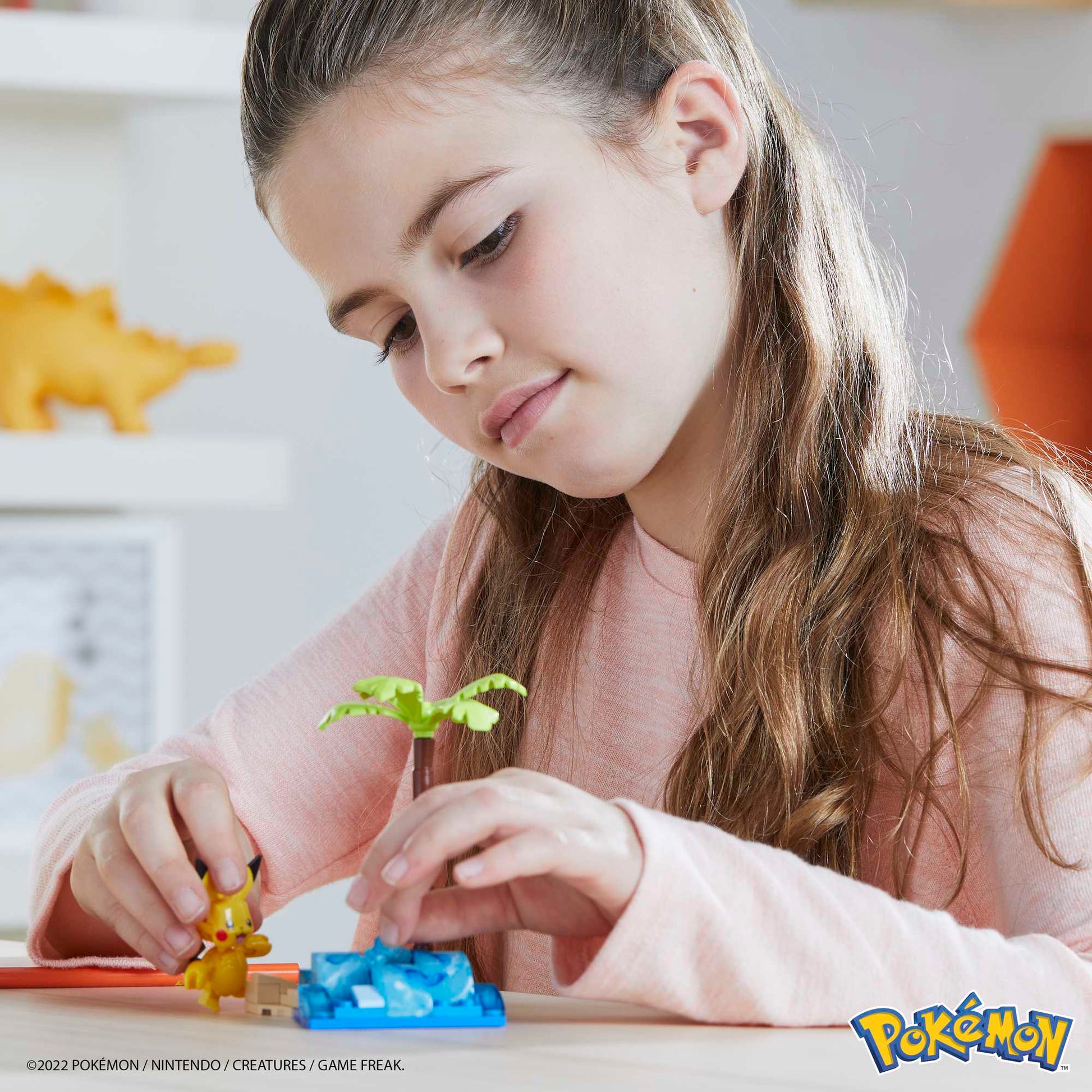 A young girl with long brown hair, wearing a light pink long-sleeve shirt, plays with the MEGA Pokemon Pikachu's Beach Splash building set by Mega Bloks. The set features a Pikachu figure, palm tree, and blue base. Pokémon logo appears at bottom right.