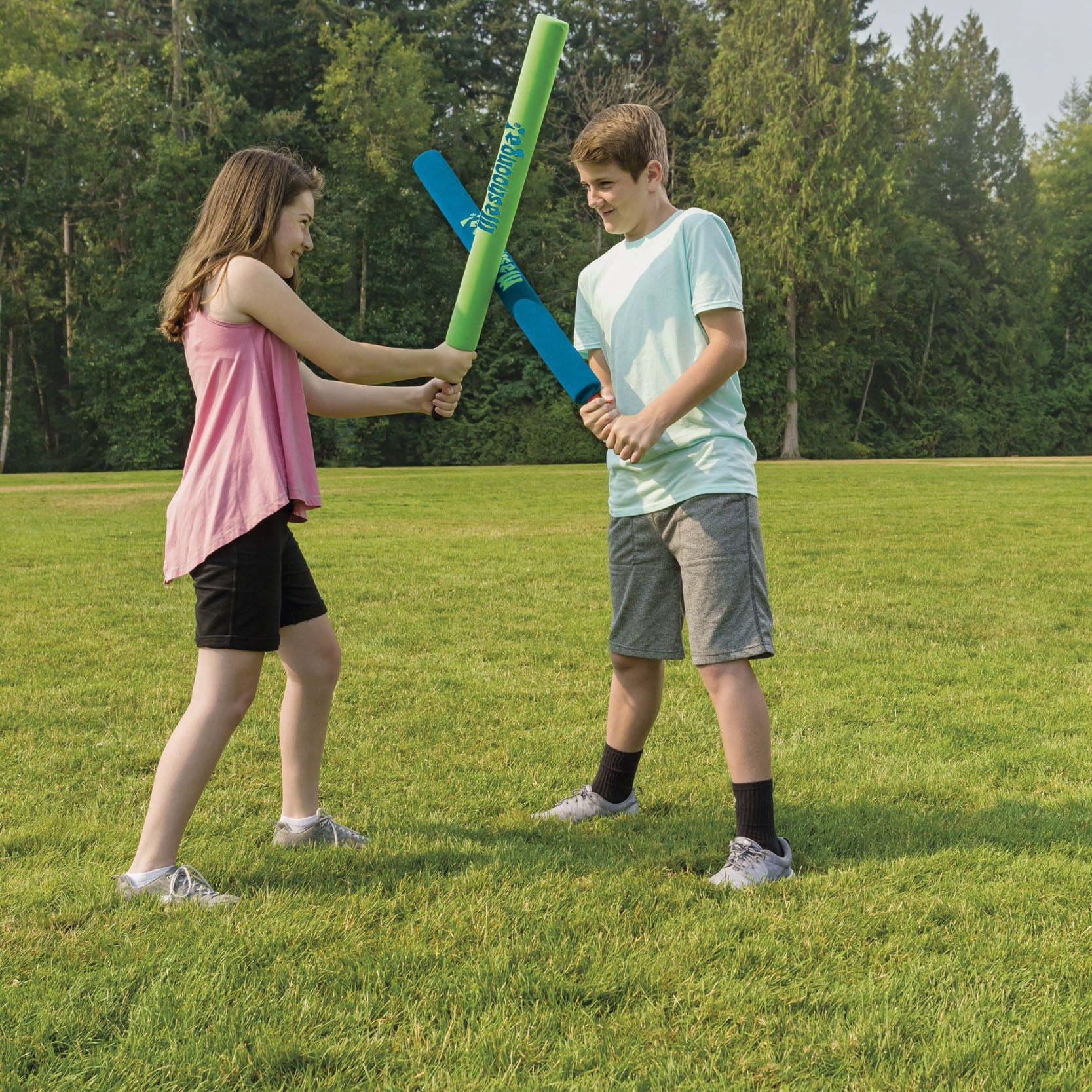 Two kids have fun on the grass, smiling as they duel with GO! Play Mashoonga foam bats from GO! Play. The girl wears a pink top and black shorts; the boy is in a light blue shirt and gray shorts. Trees are visible in the background.