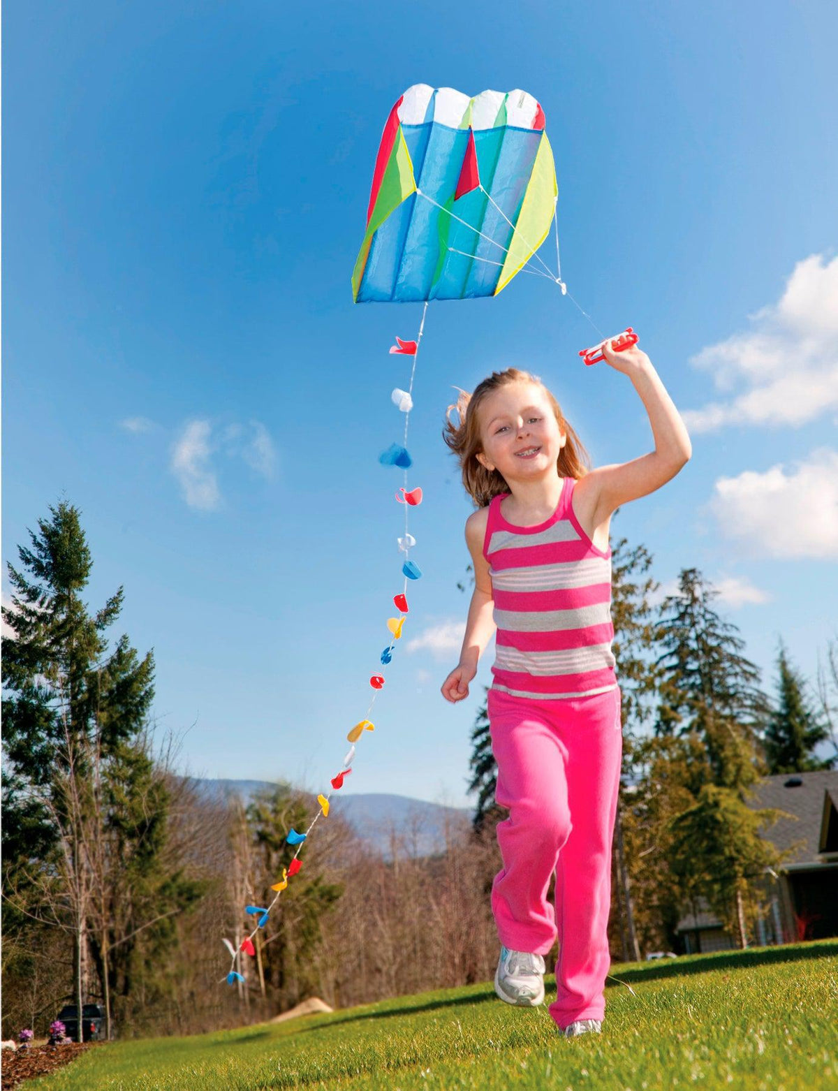 A young girl in pink runs on grass, enjoying outdoor fun as she flies the GO! Launch Parafoil Kite by GO! Launch under a bright blue sky with trees and a house in the background.